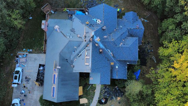 An aerial view of a house undergoing roof repairs, featuring exposed wood decking, stacks of shingles, and a dumpster.