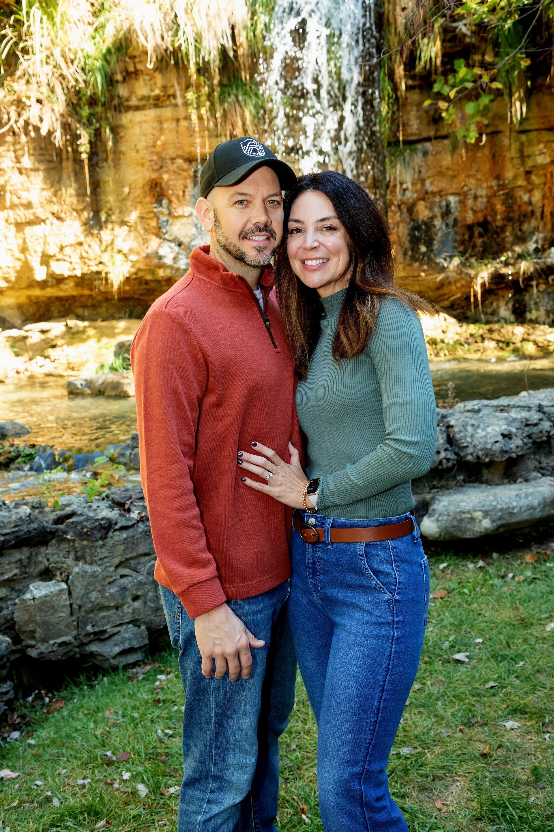 A smiling couple stands outdoors in front of a waterfall, the person on the left wearing an orange sweater and ball cap.