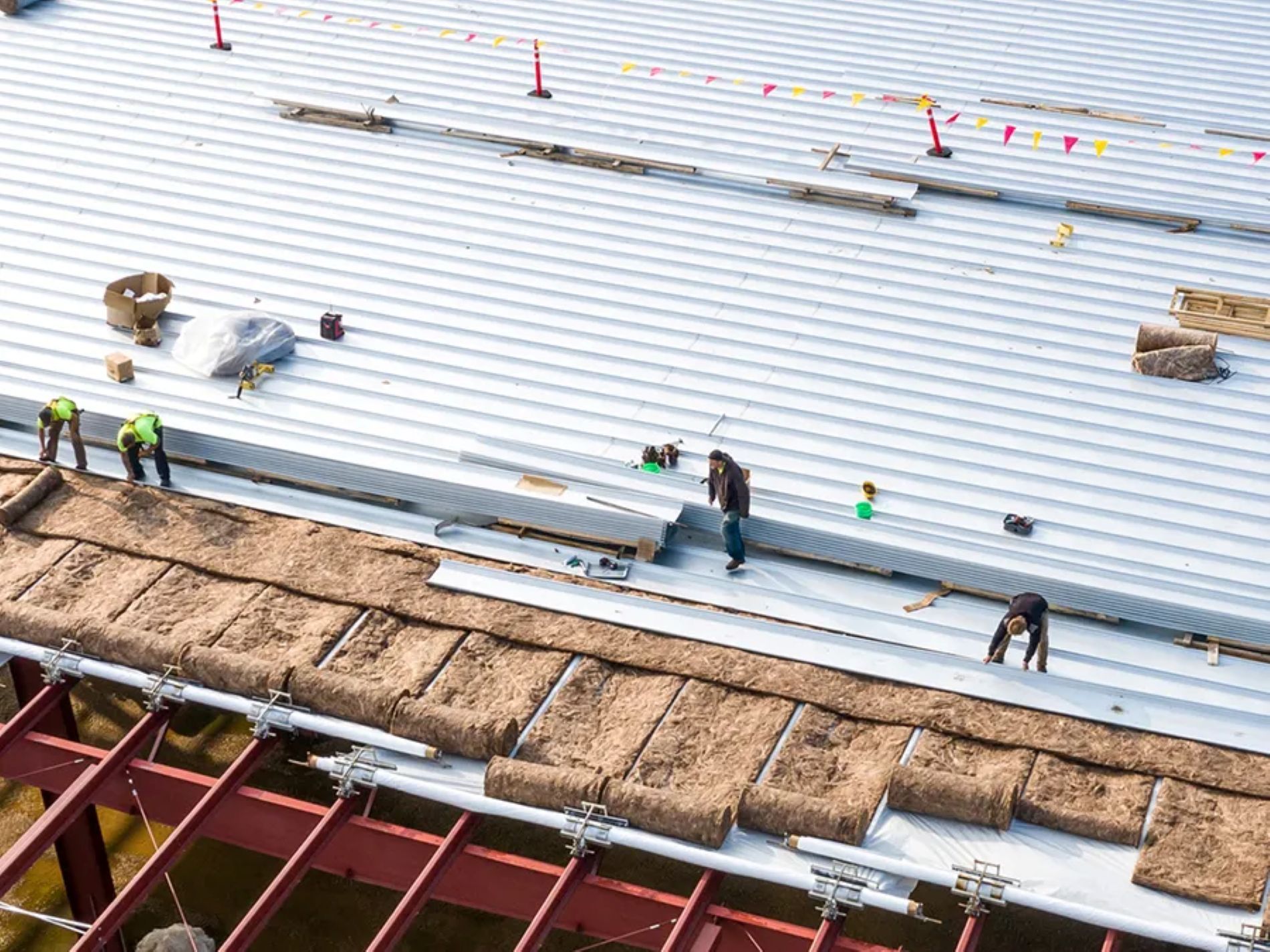 Construction workers in high-visibility vests install insulation and roofing panels on a large steel-framed structure.