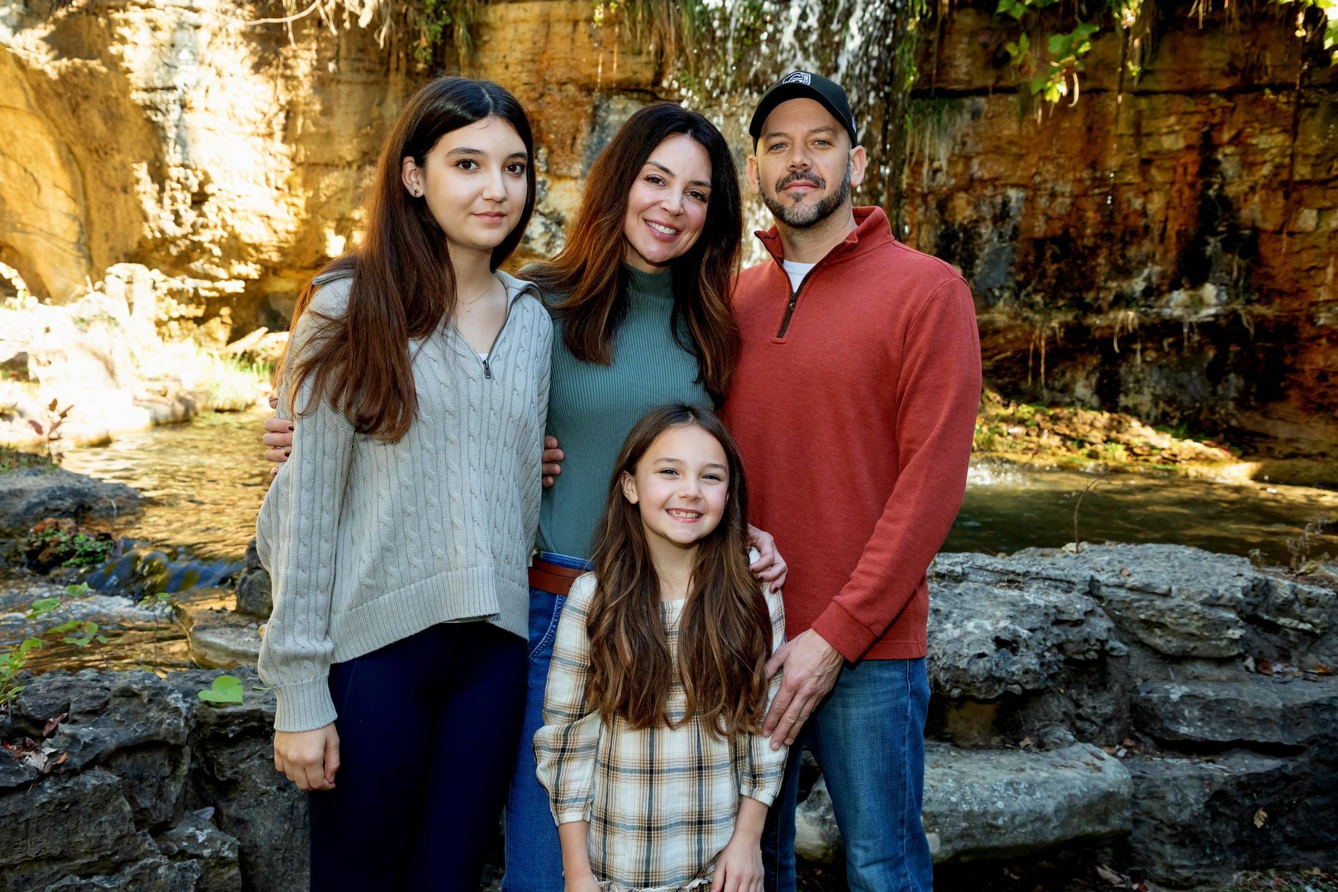 A family poses for a portrait outdoors in front of a rocky ledge and greenery, wearing casual sweaters and a jacket.