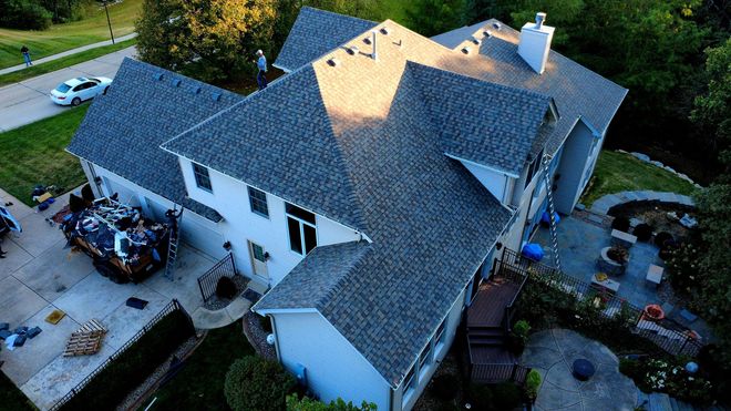 An aerial view of a multi-story house with a grey shingled roof, showing an adjacent driveway and a backyard patio area.