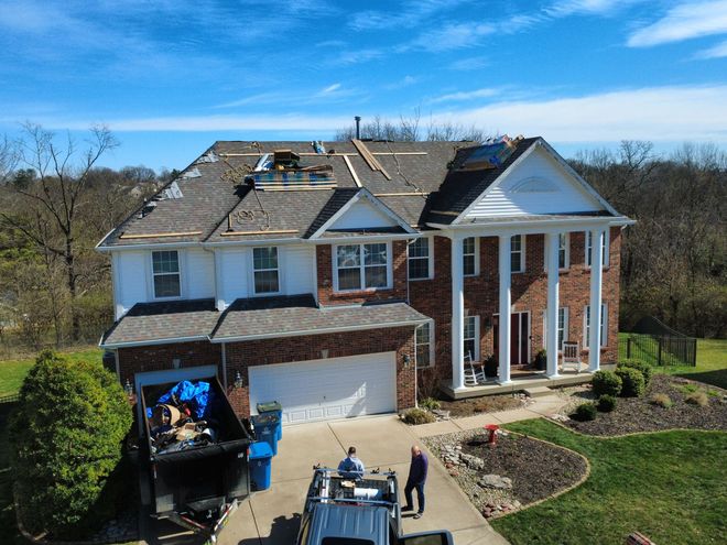 Workers repairing the roof of a two-story brick colonial home with a dumpster in the driveway.