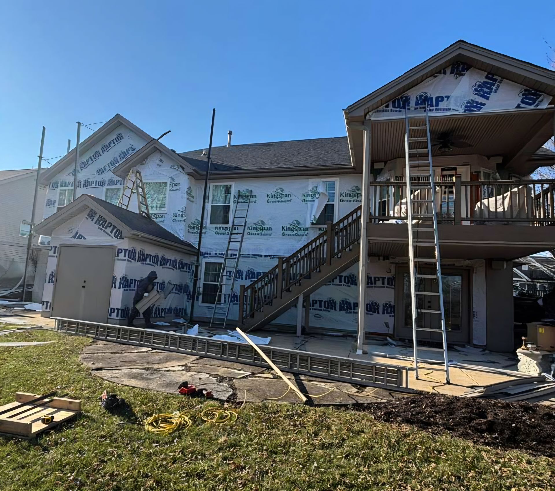 Partially built two-story house under construction with stairs, scaffolding, and house wrap in a sunny yard