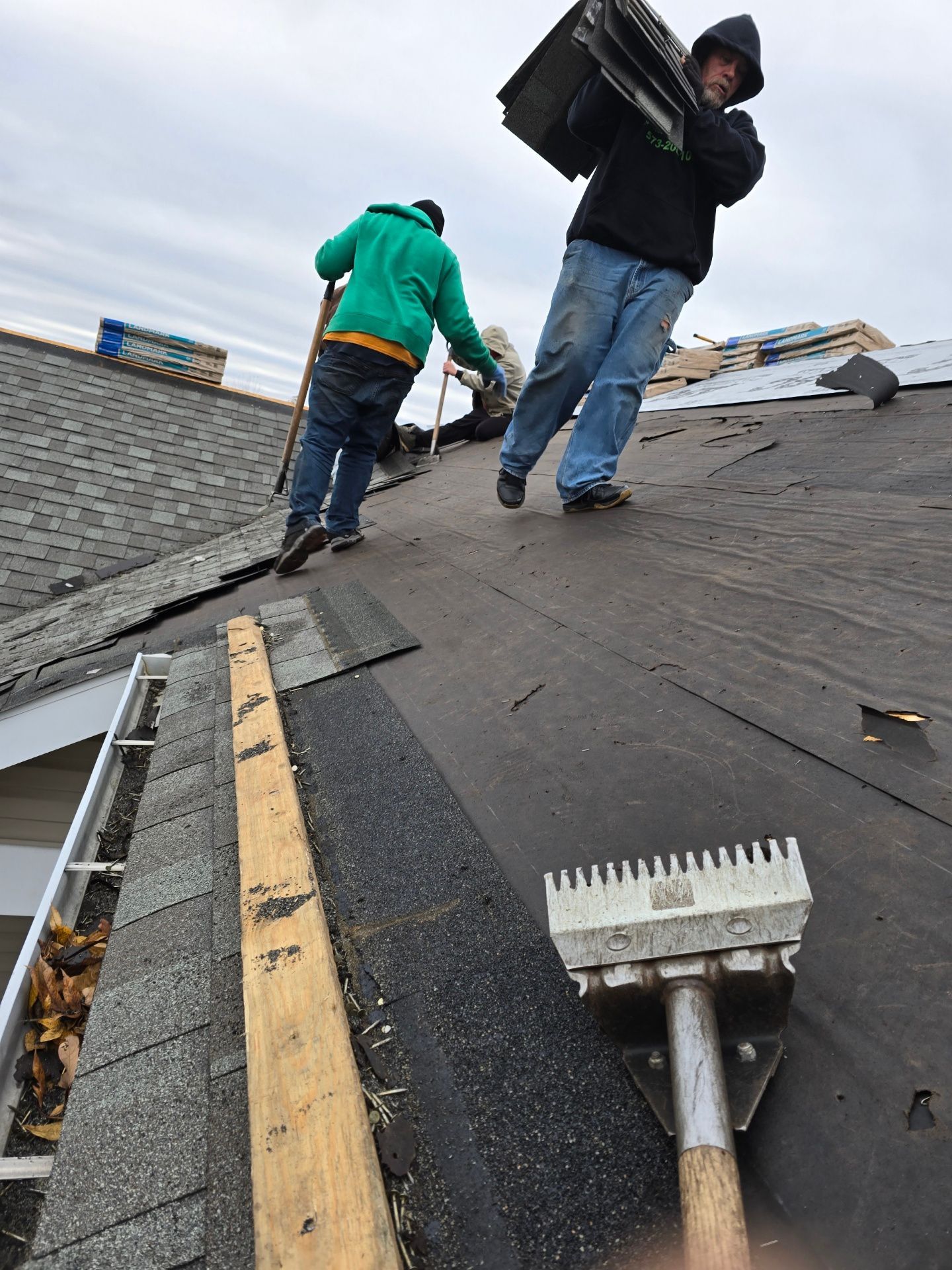 Two construction workers in hoodies remove shingles from a residential roof, with a long-handled scraper in the foreground.