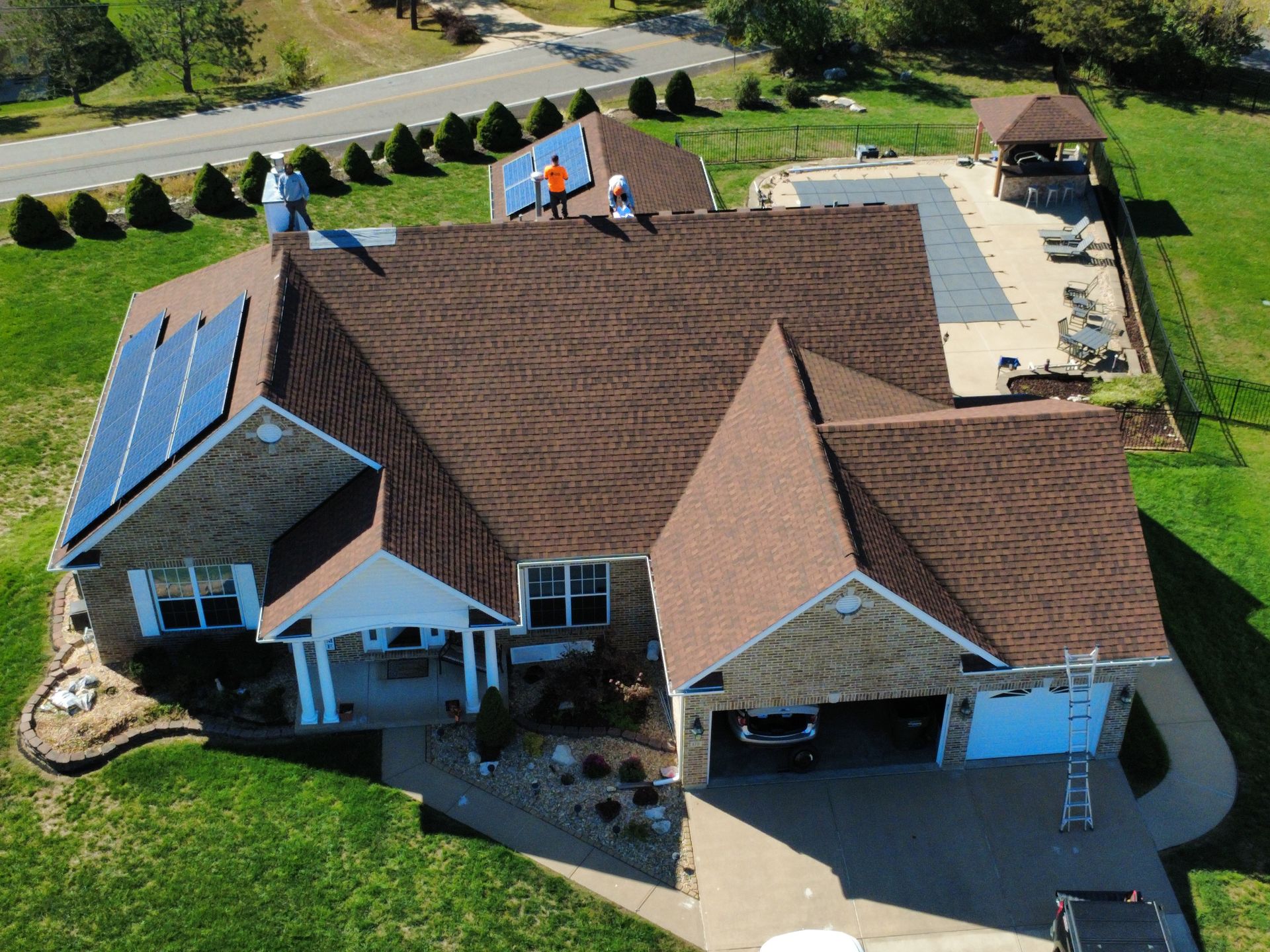Aerial view of a brown shingled home with solar panels, construction workers on the roof, and a pool in the backyard.