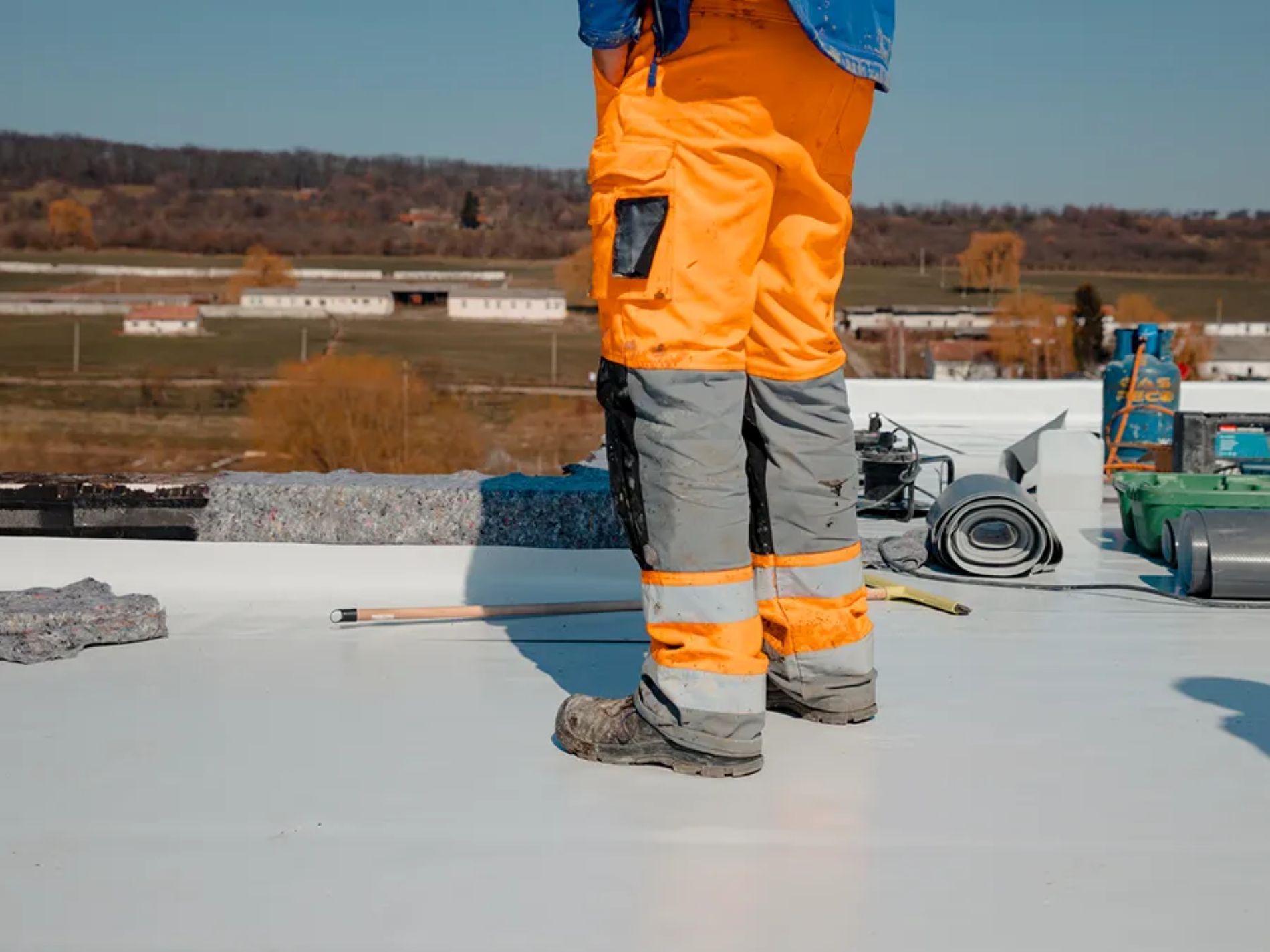 A worker in high-visibility orange and grey work pants stands on a white rooftop under a clear blue sky.