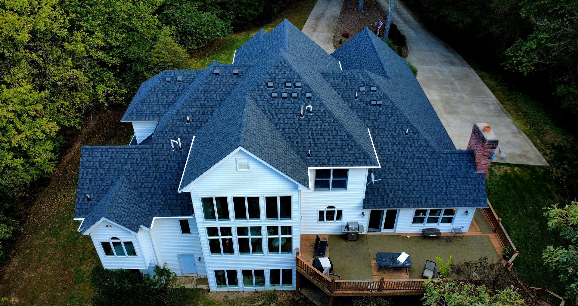 Aerial view of a white two-story house with a dark roof, large windows, and a back deck, surrounded by trees.