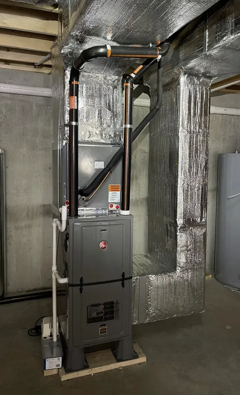 Gray HVAC furnace in a basement with insulated ductwork and metal piping.