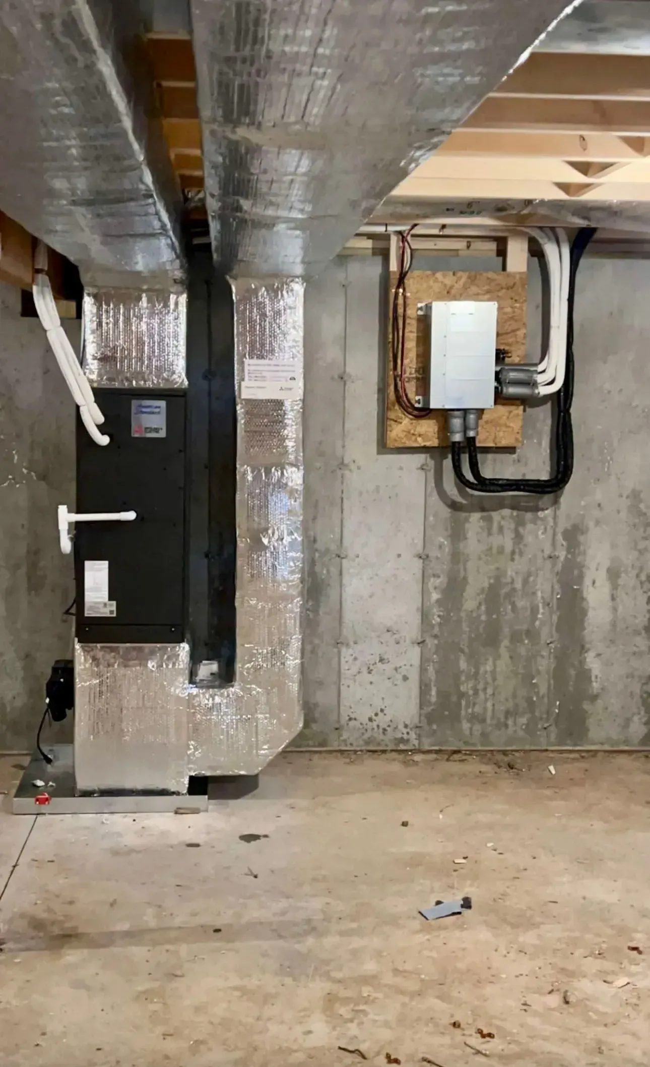 Basement utility area with exposed silver ductwork, pipes, and a wall-mounted electrical panel on concrete walls.