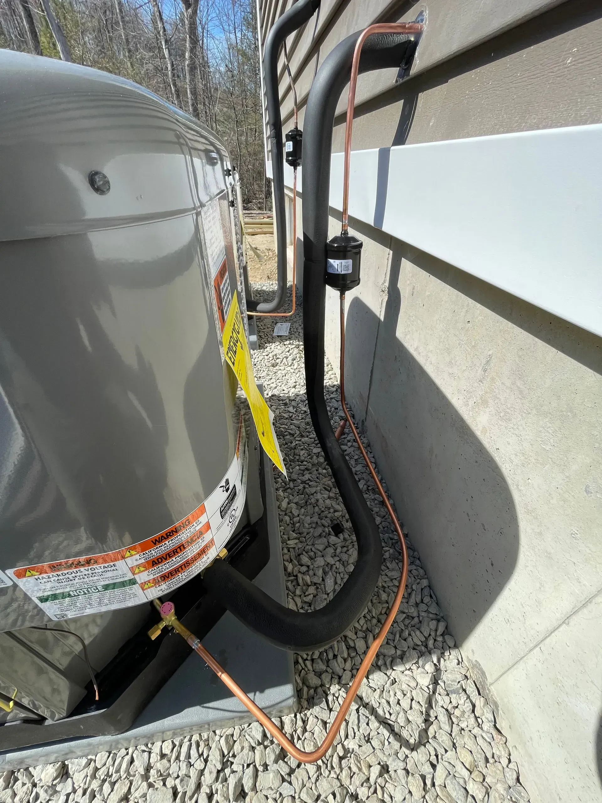 Outdoor HVAC unit beside a house, with black insulated tubing and orange wires on gravel.