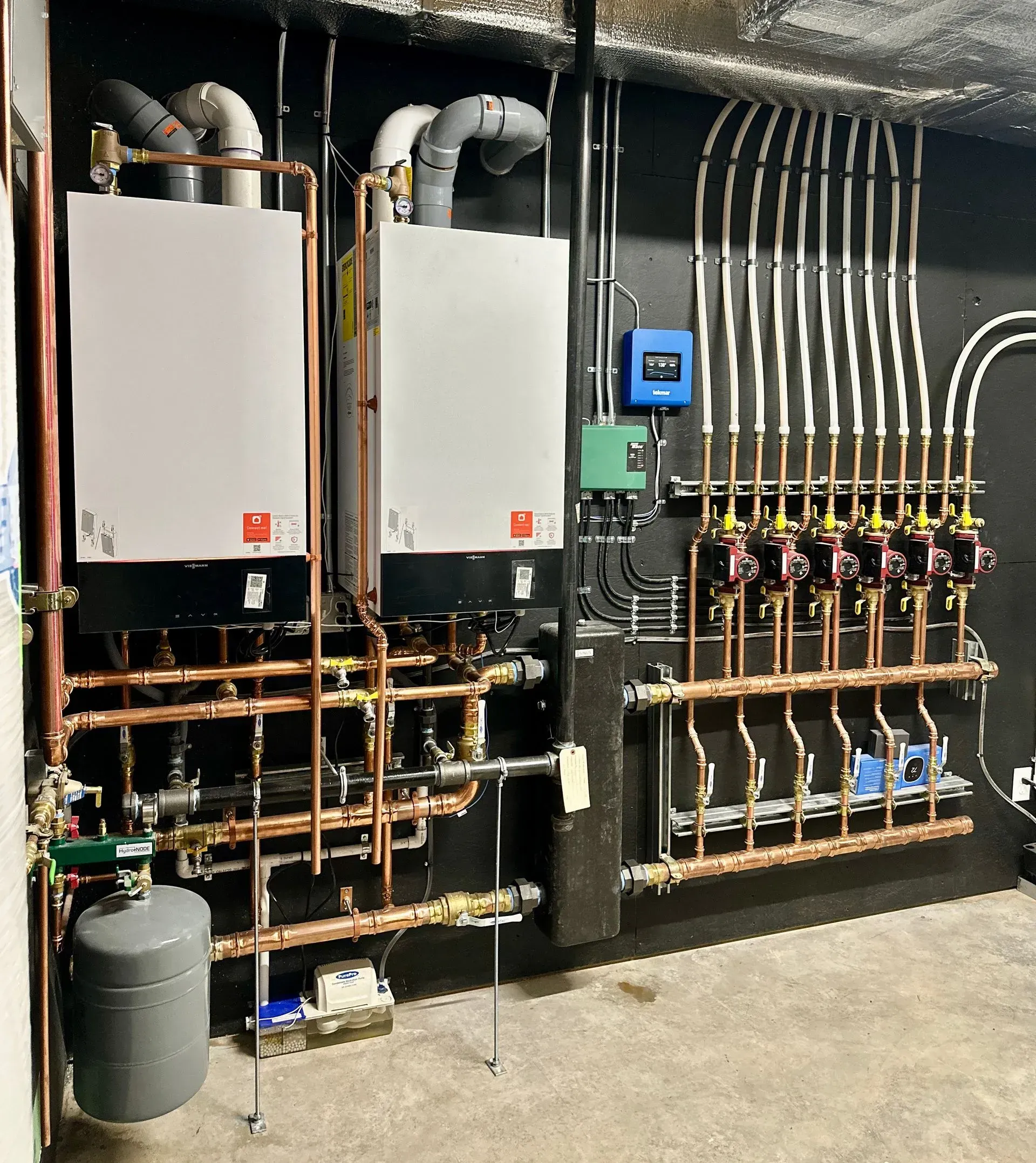 Boiler room with two white wall-mounted units, pipes, gauges, and control manifolds on a black wall.