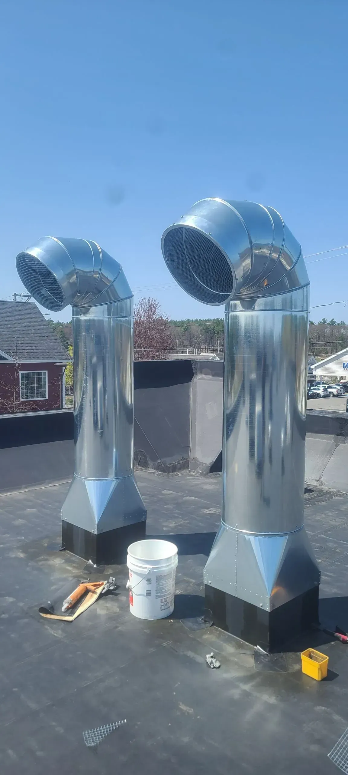 Two silver rooftop ventilation ducts on a flat roof beside a white bucket and tools.