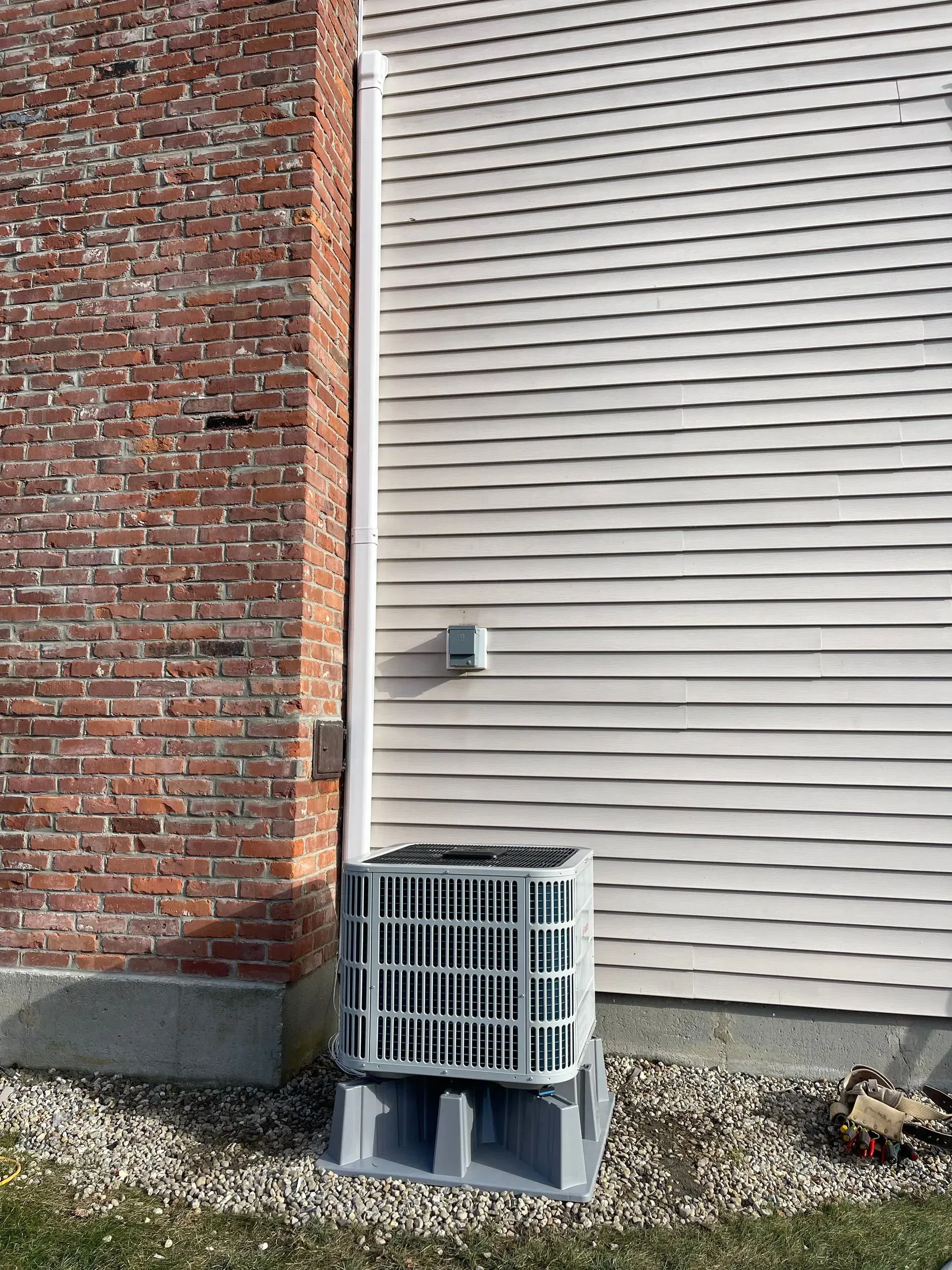 Outdoor air conditioner unit beside a brick wall and beige siding on a gravel base