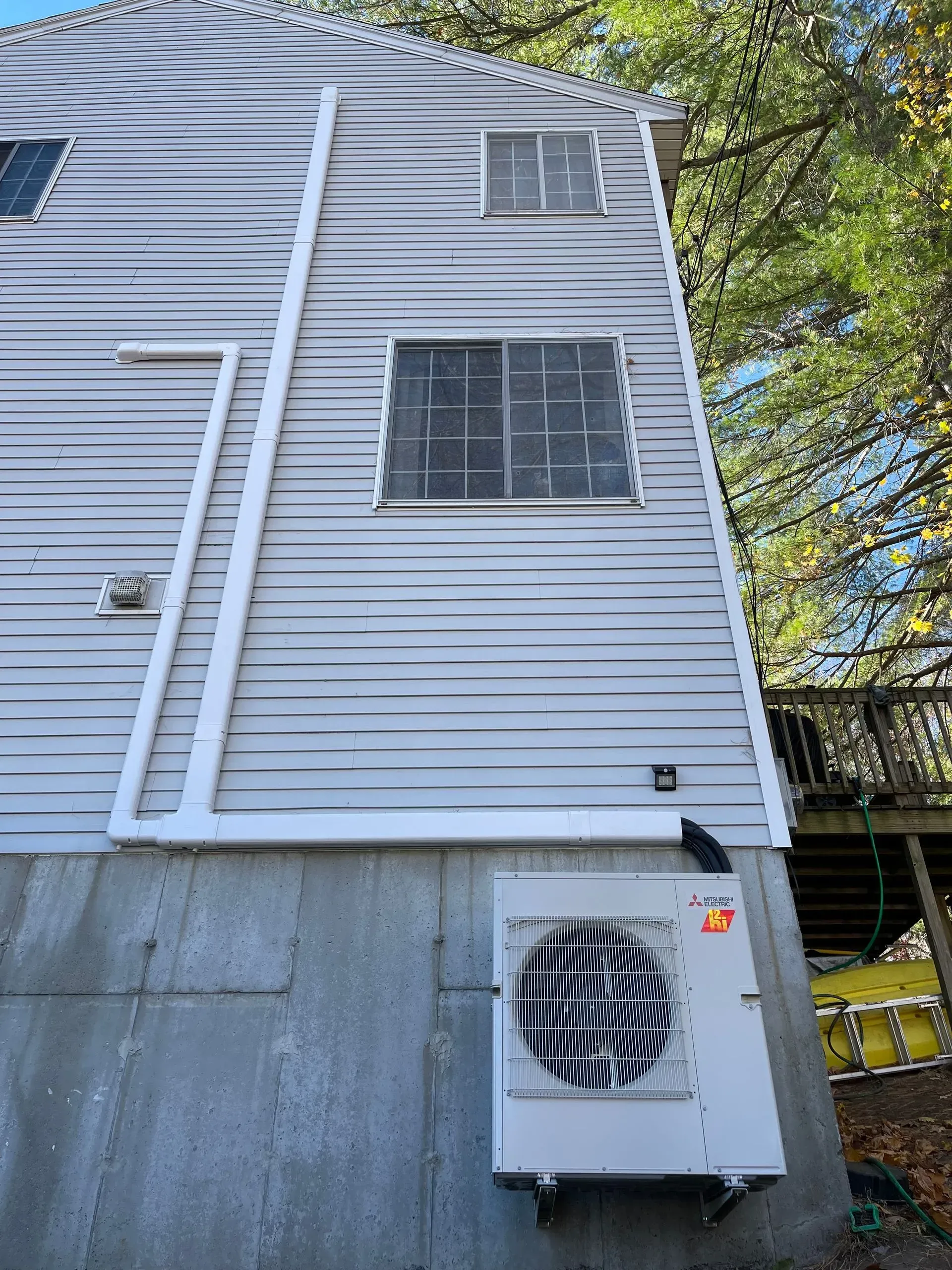 Side of a gray building with windows, vertical pipes, and an outdoor HVAC unit on a concrete foundation