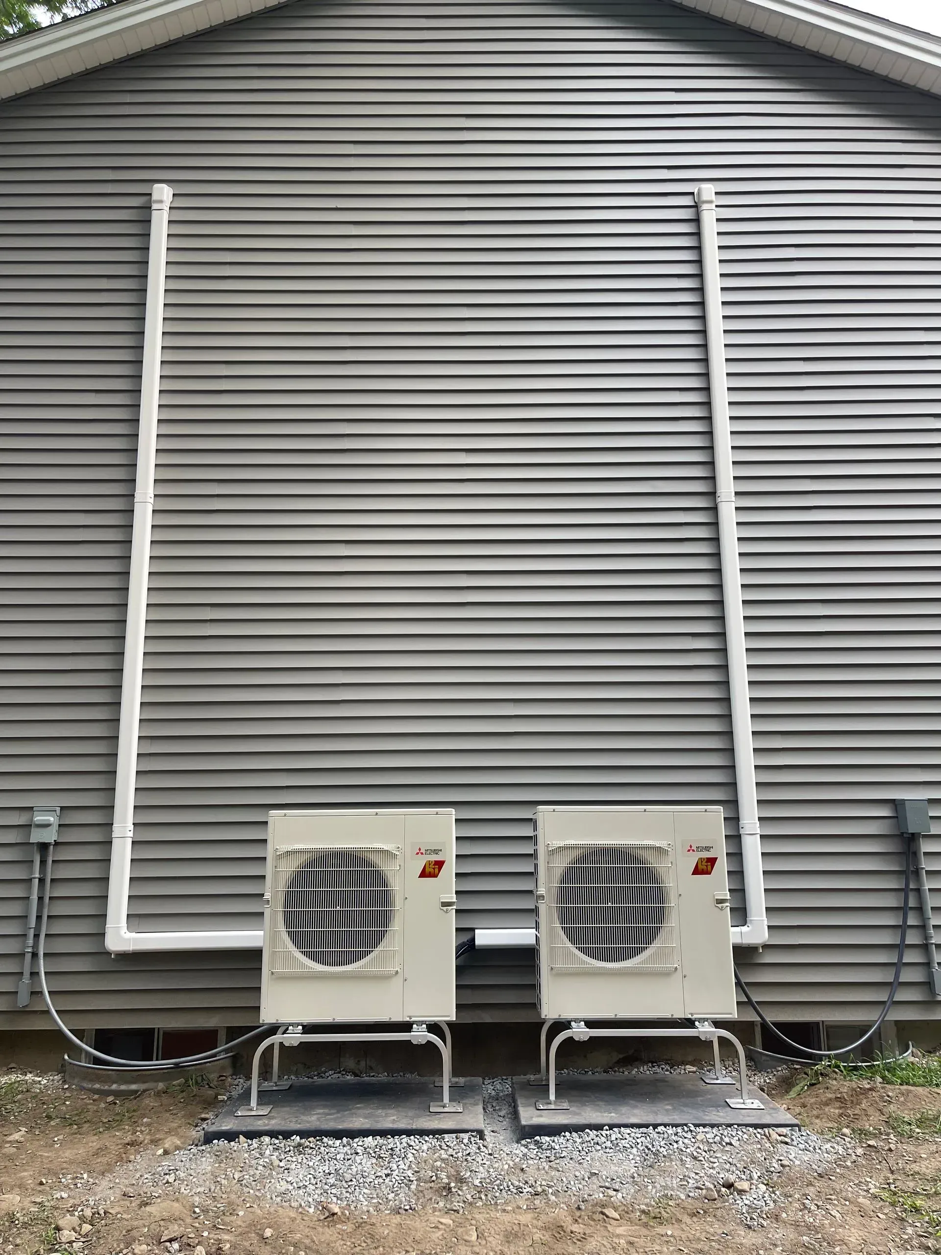 Two outdoor HVAC units on gravel beside a gray building wall with vertical pipes