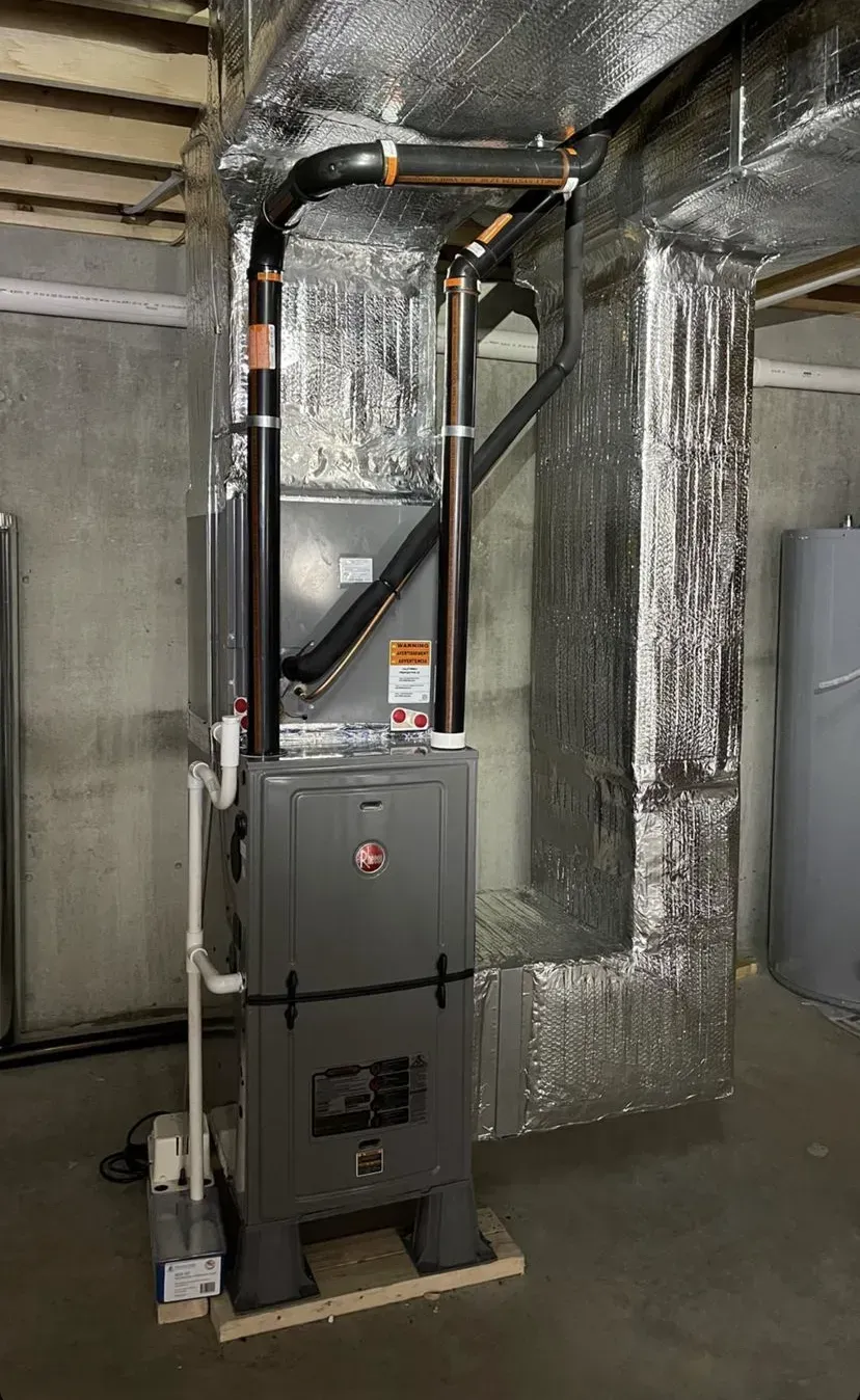 Gray furnace in a basement utility area with insulated ducts and exposed pipes.