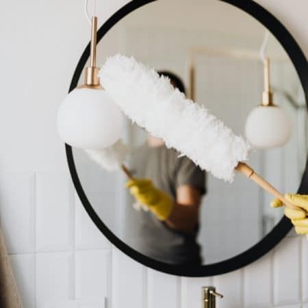 Person dusting a round mirror with a white duster in a bathroom, wearing yellow gloves.