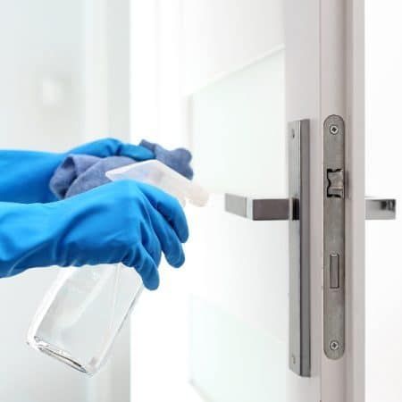 Person in blue gloves sanitizing a white door handle with spray and cloth.