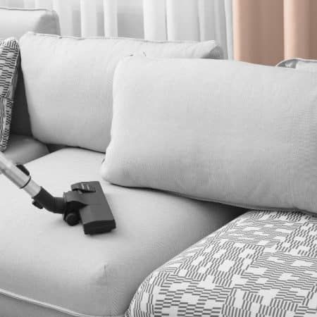 Vacuuming a light gray couch with a black vacuum cleaner head.