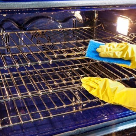 Person in yellow gloves cleaning oven racks with a blue sponge.