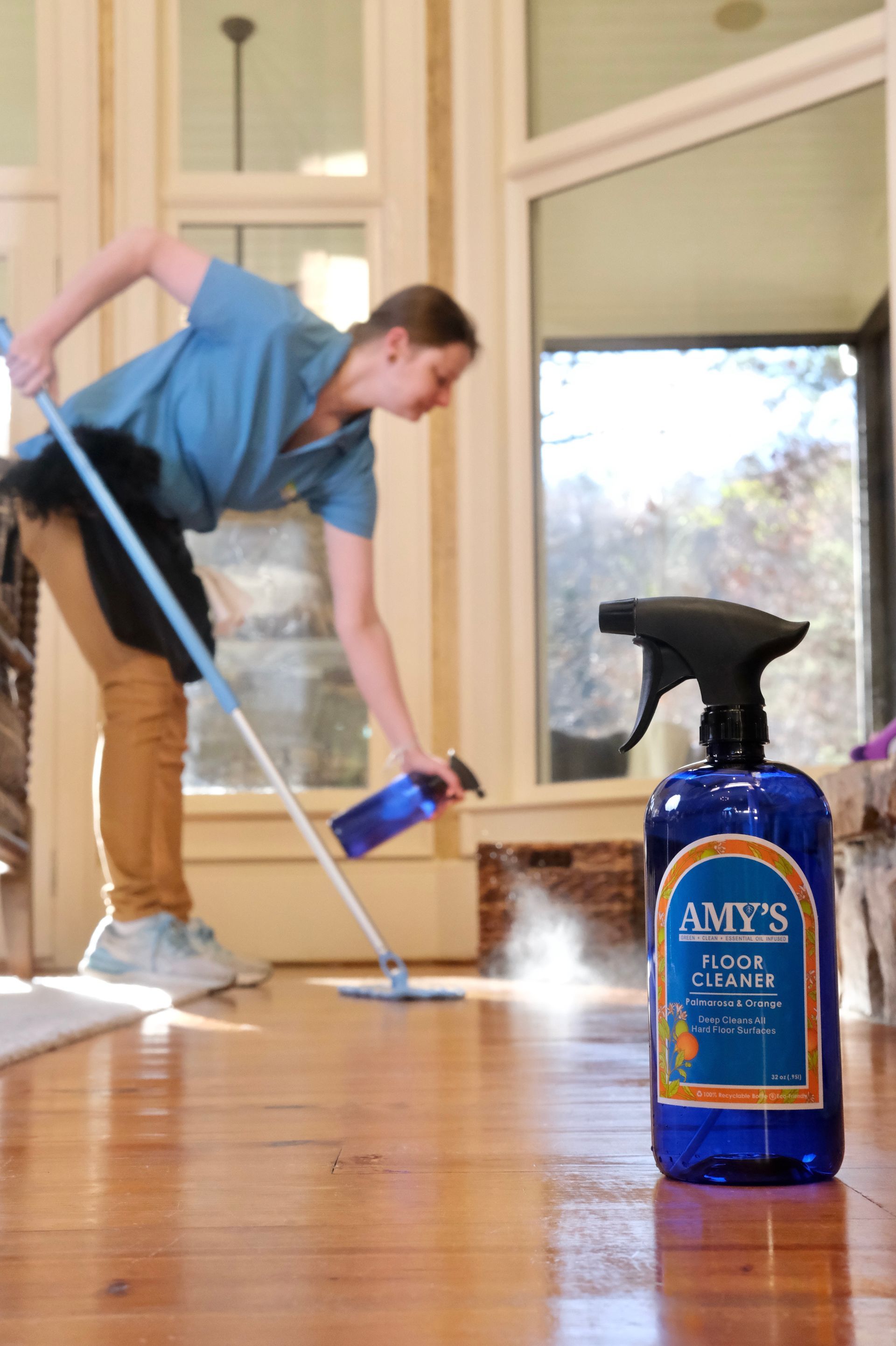 Woman mopping wood floor, spraying cleaner. Blue cleaner bottle in foreground. Bright room.
