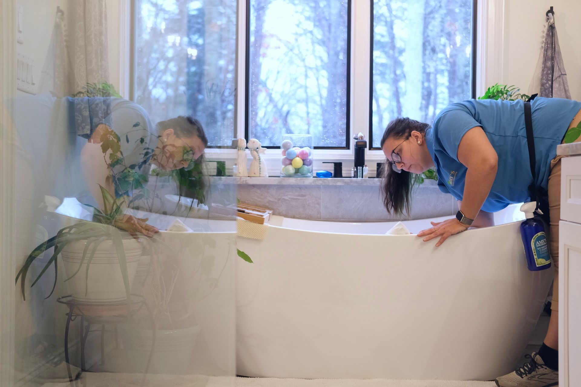 Woman leans over a bathtub, inspecting the water. Large window in background.