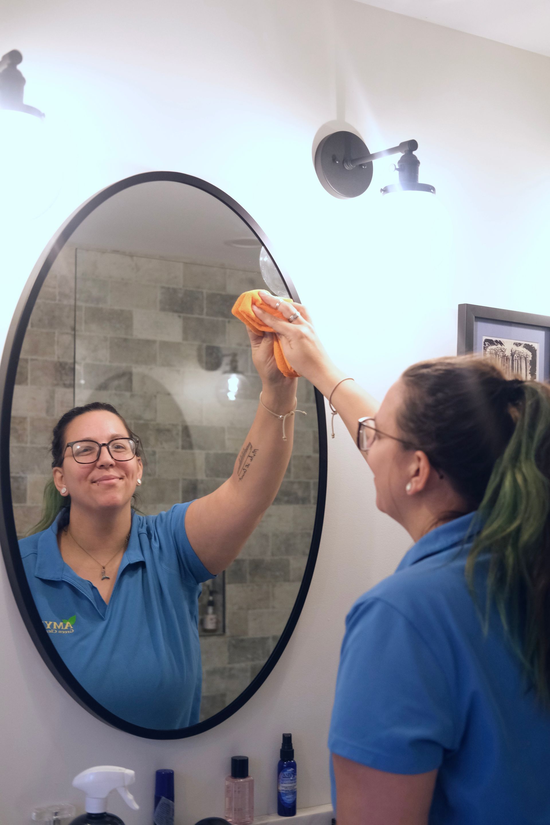 Woman in blue shirt cleans oval mirror in bathroom.