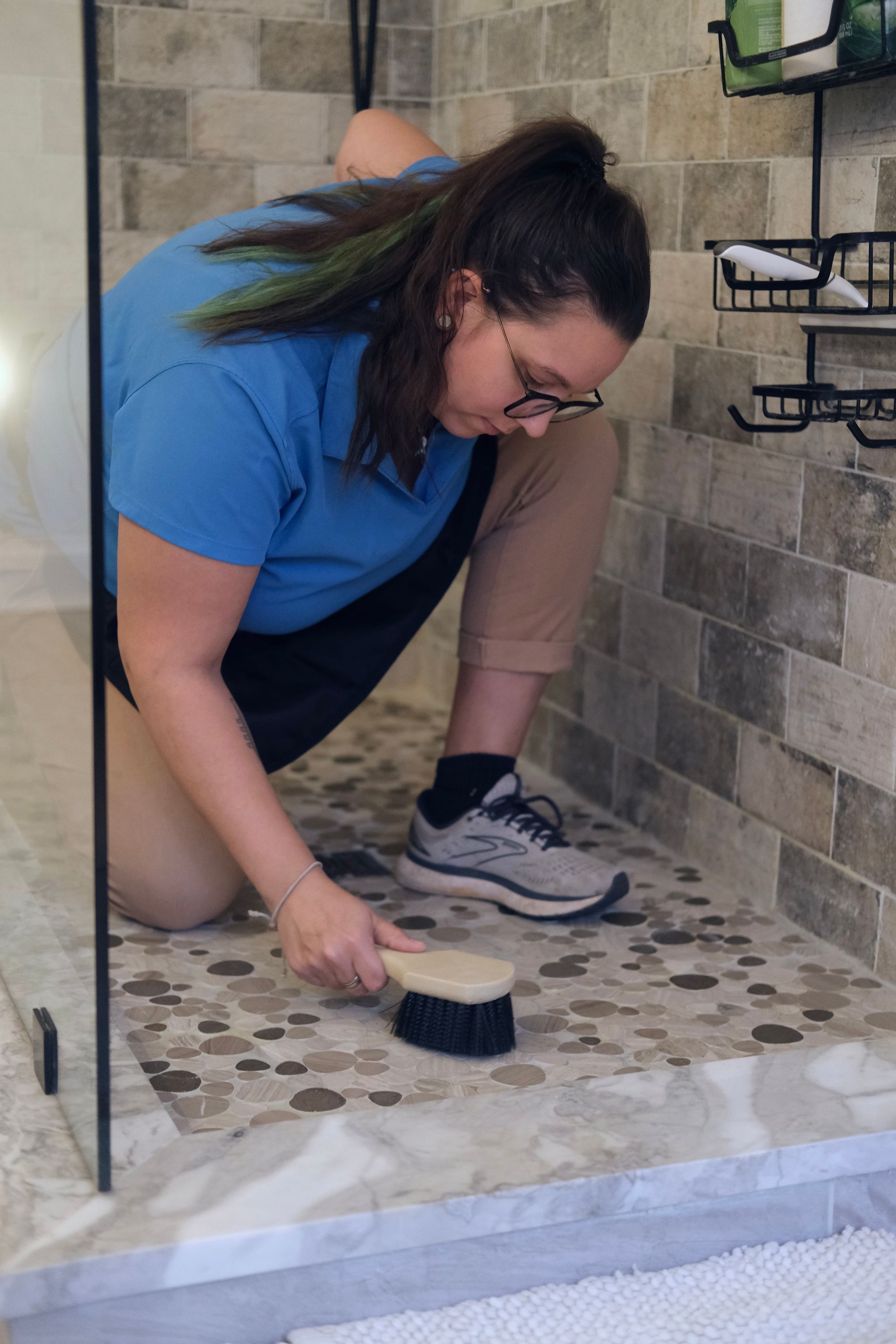Woman in blue shirt scrubs shower floor with brush.