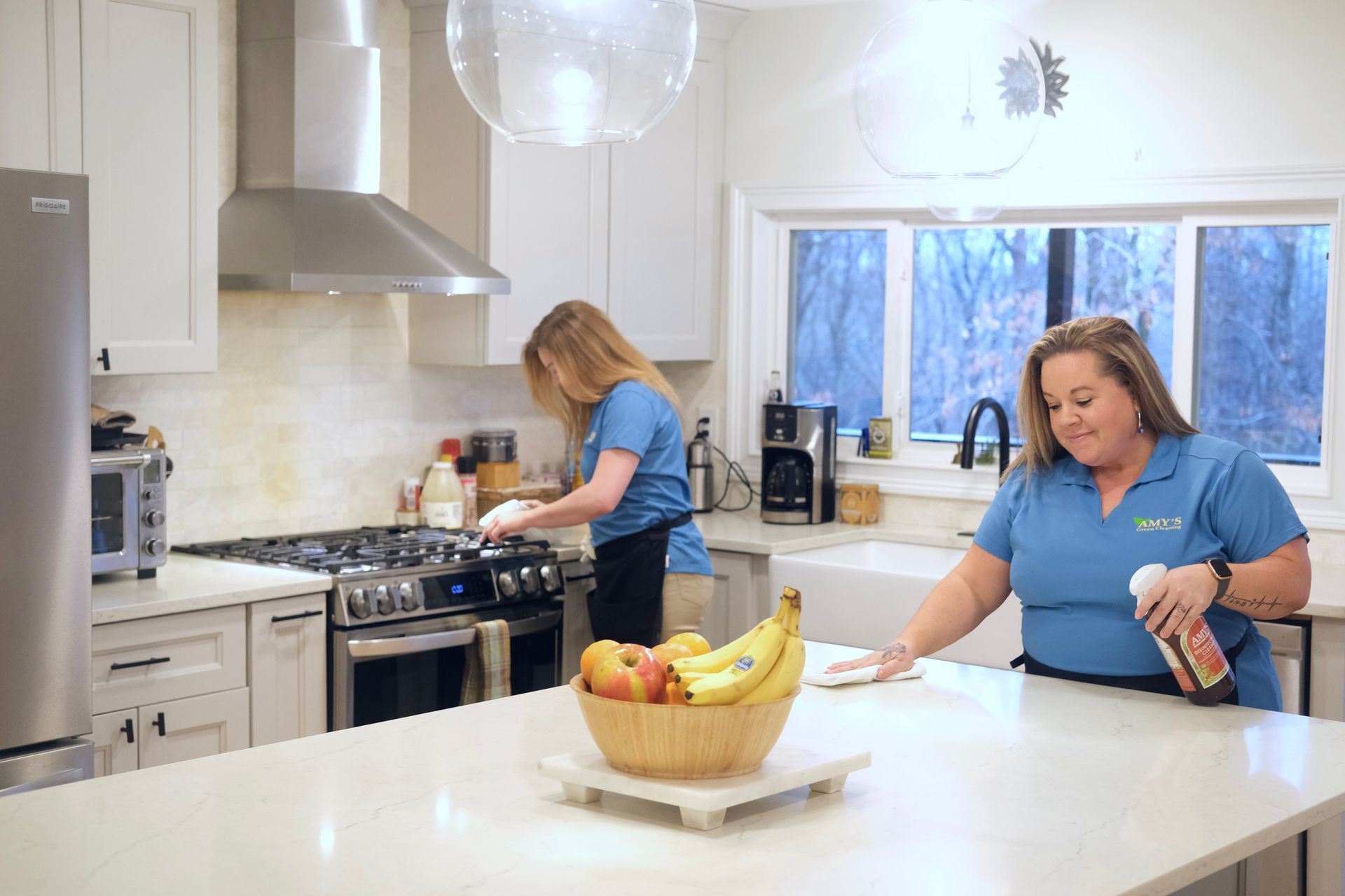 Two women wearing blue cleaning shirts clean a bright kitchen. One cleans the stovetop; the other, the countertop.