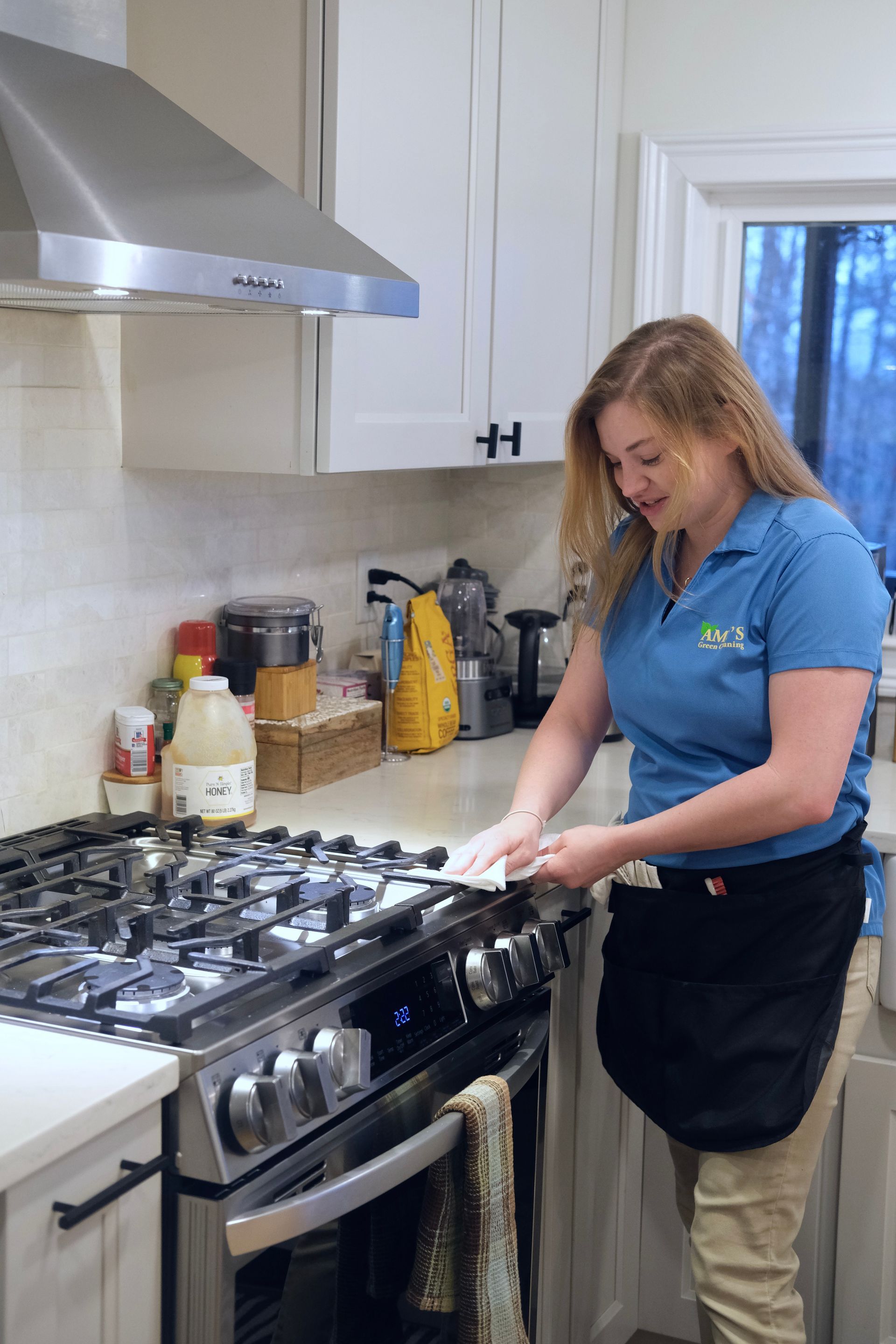 Woman in blue shirt cleaning a stainless steel gas stove in a kitchen.