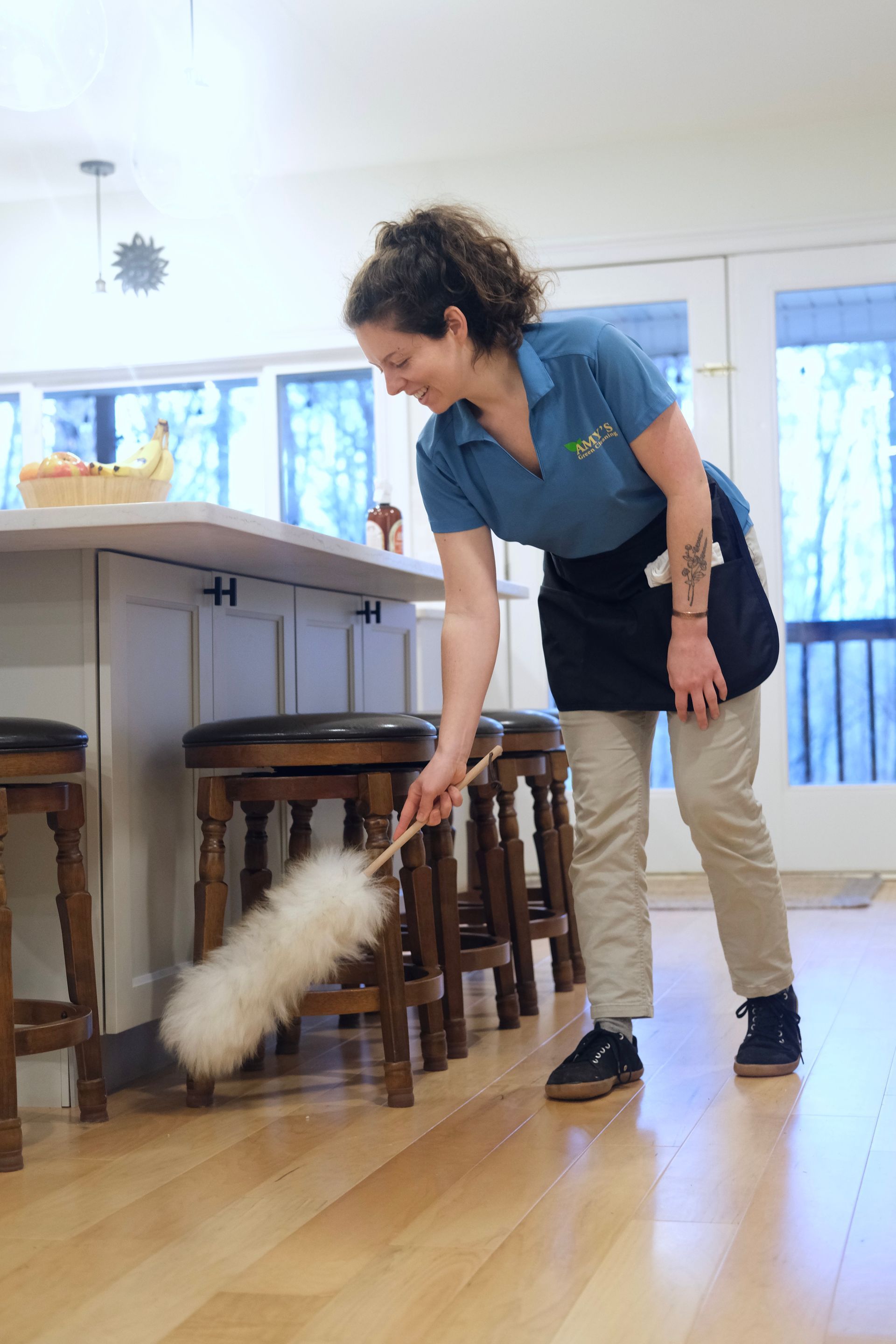 Woman dusting stools in a kitchen, smiling. She wears a blue shirt, apron, and tan pants.