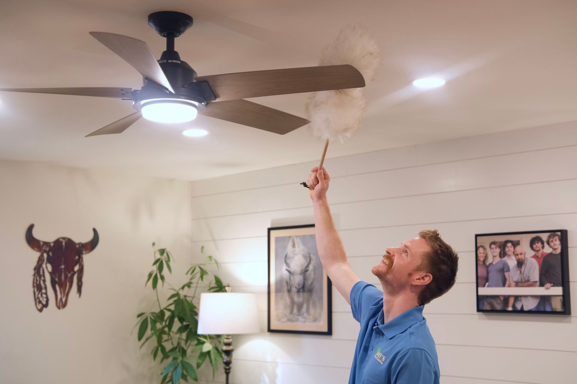 Man dusts a ceiling fan in a brightly lit room. He has a feather duster and smiles, looking up.