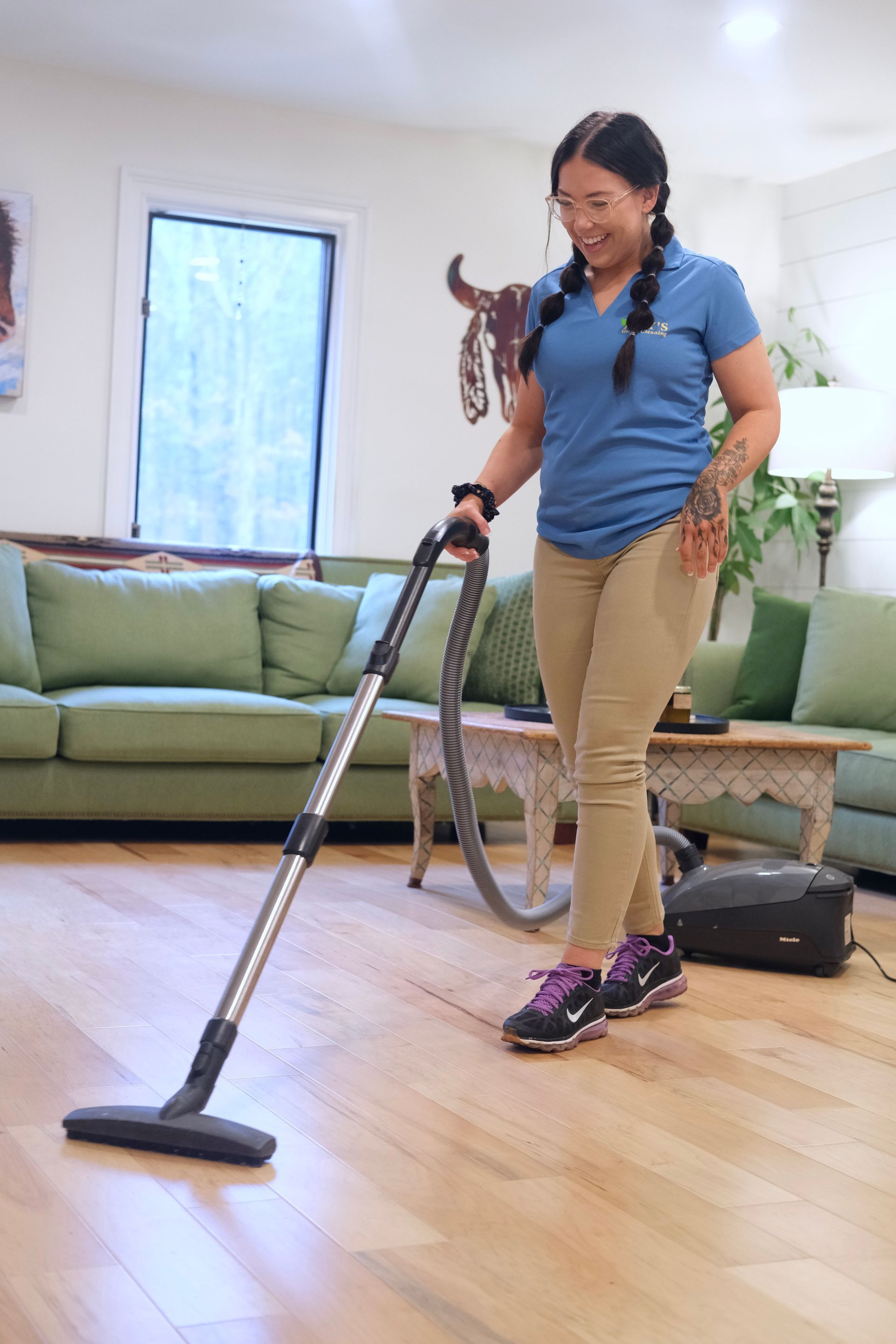 Woman with pigtails vacuums a hardwood floor in a living room with green couch and a window.