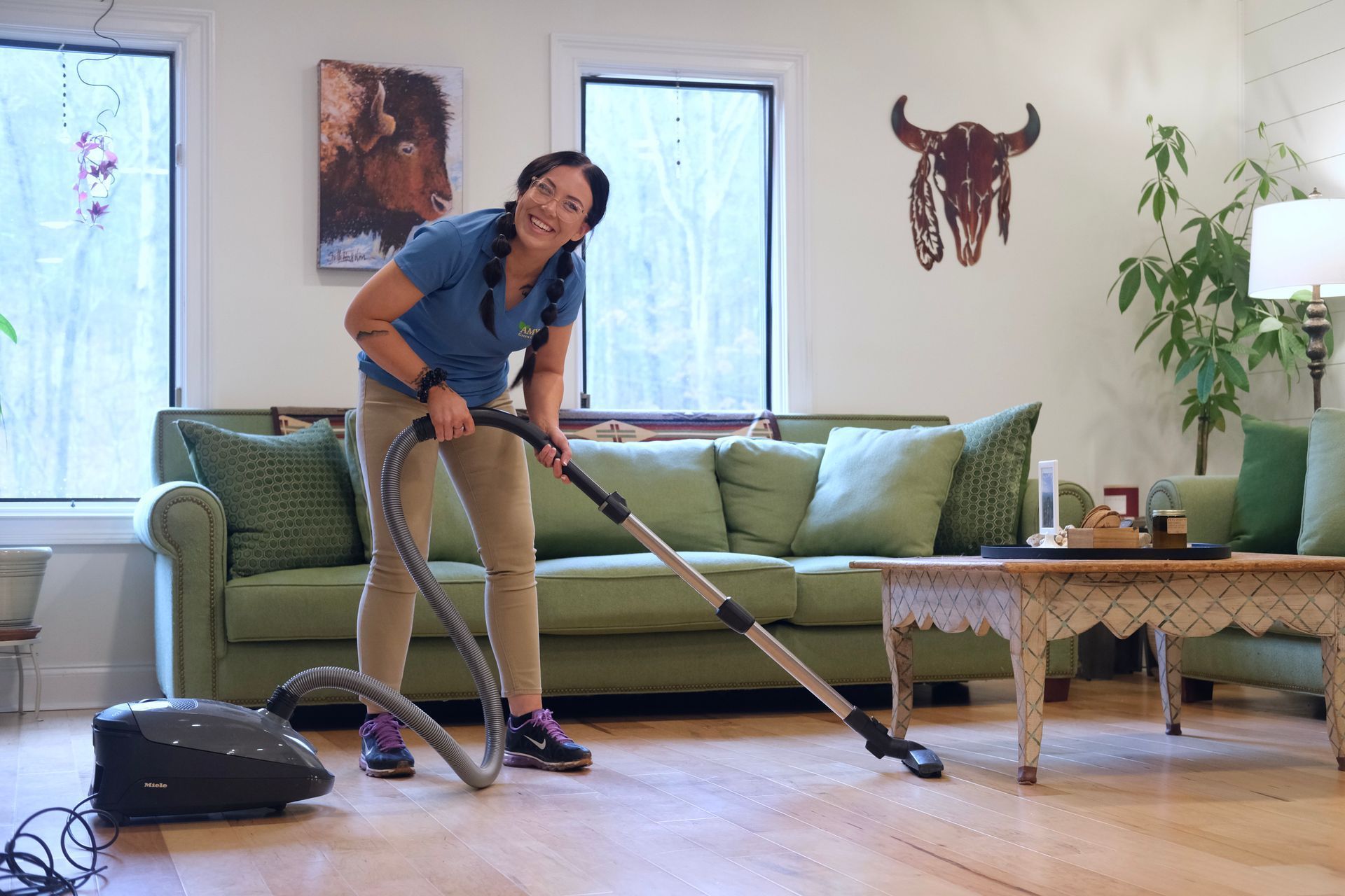 Woman vacuuming wooden floor in living room, smiling. Green sofa, artwork, and plants visible.