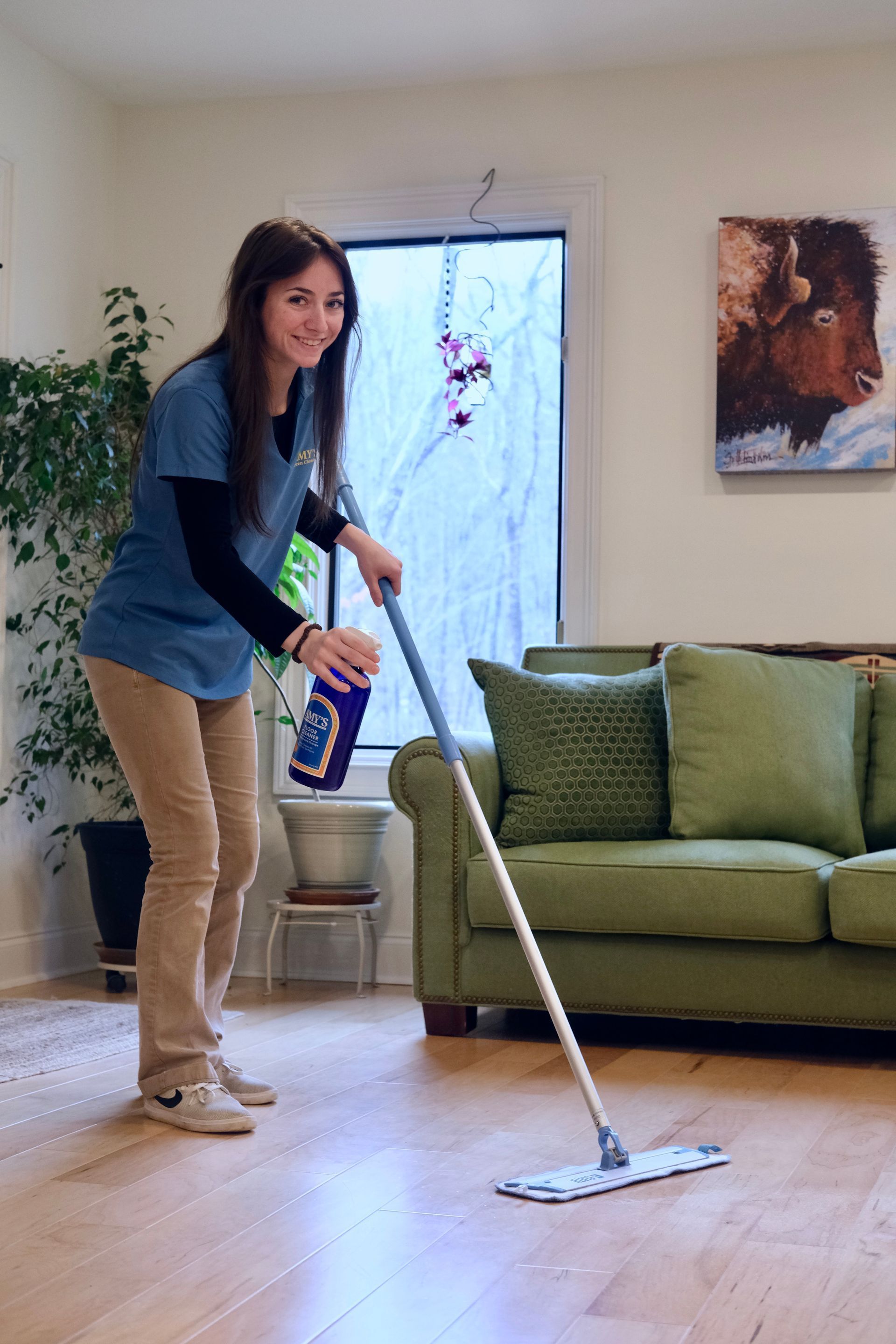Woman mopping a hardwood floor, holding a spray bottle, smiling. Living room with green couch, plants, and painting.