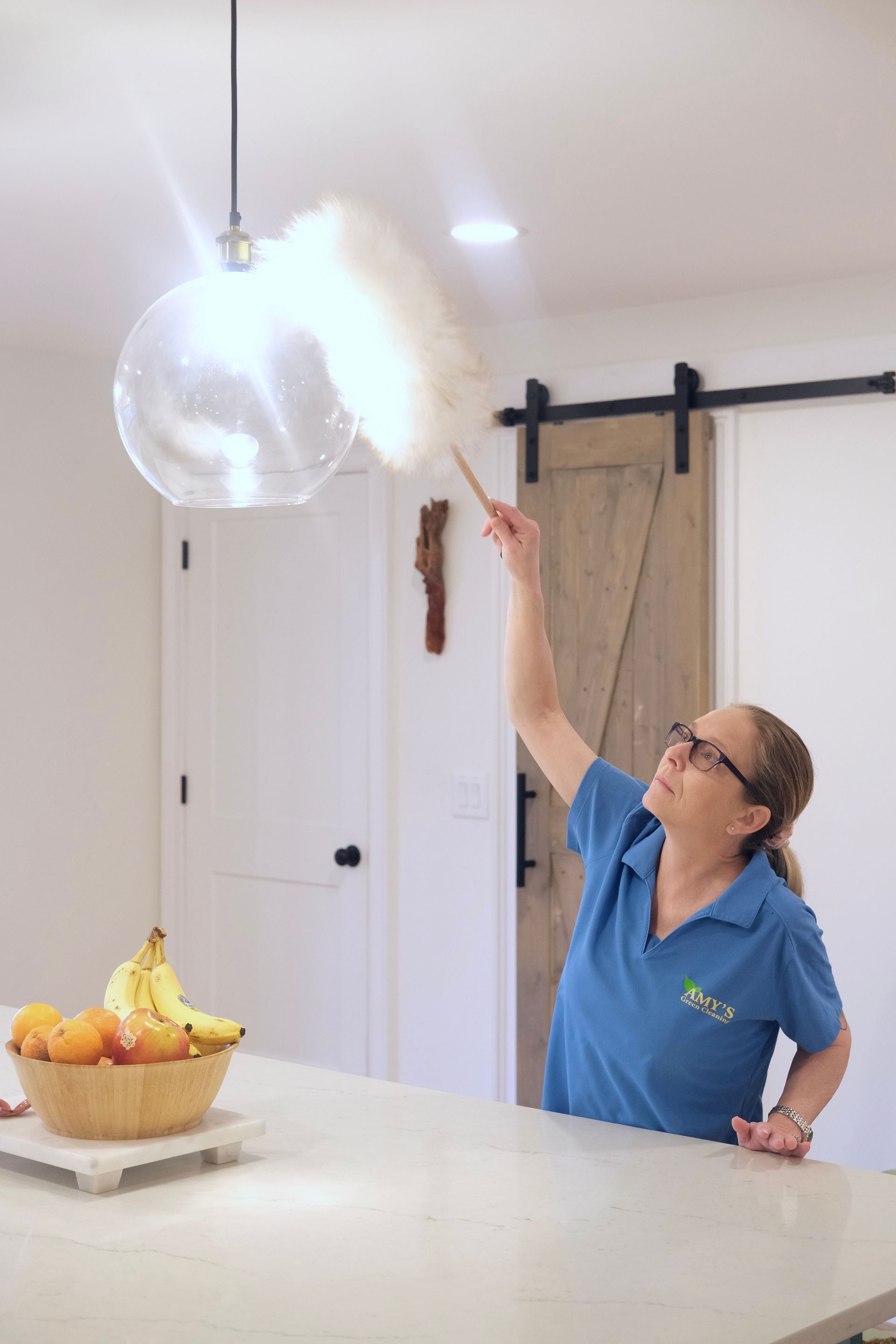 Woman dusting a glass globe light fixture with a duster in a kitchen.
