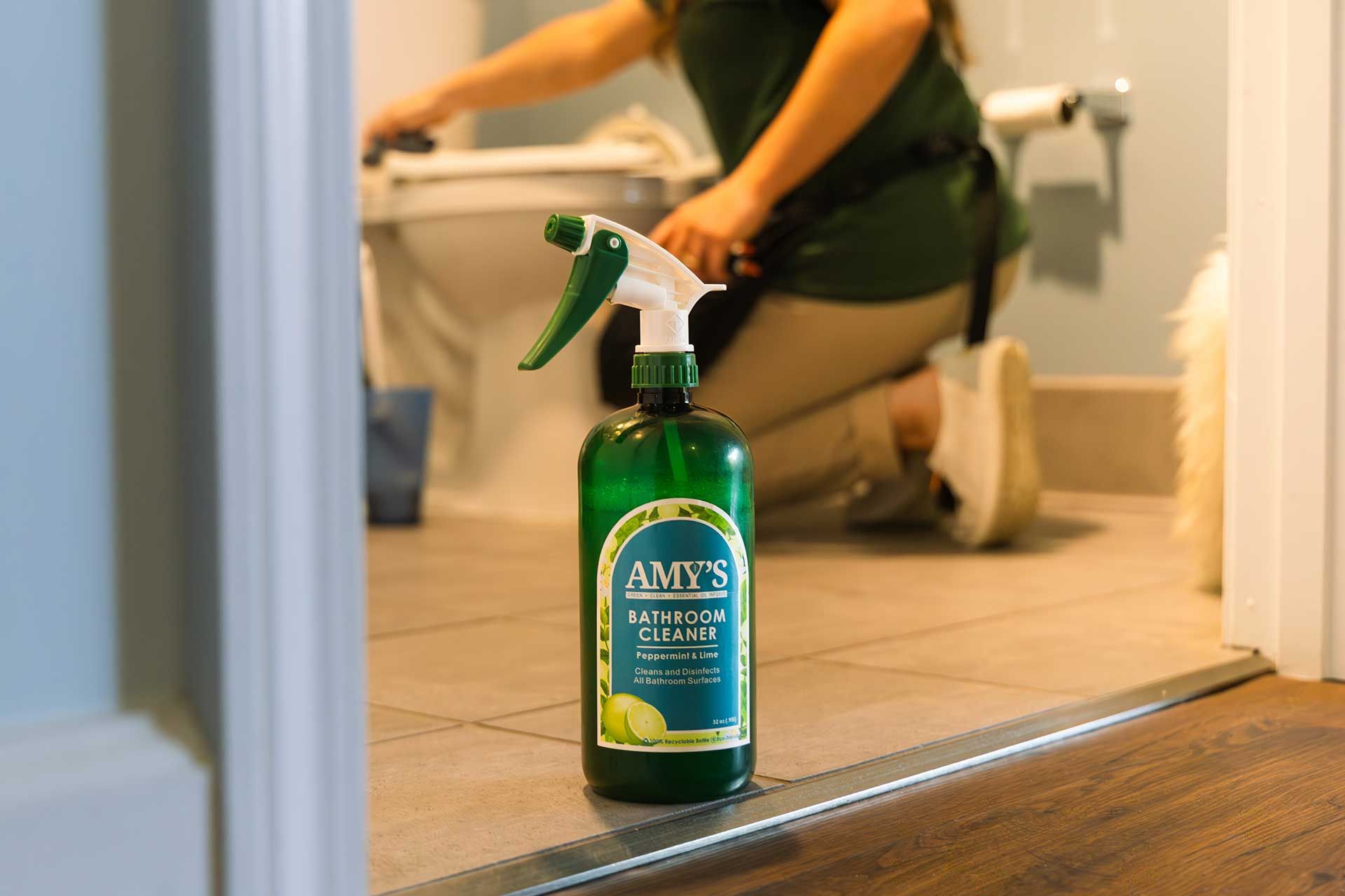 green spray bottle of toilet cleaner in front of a person cleaning a toilet in a bathroom
