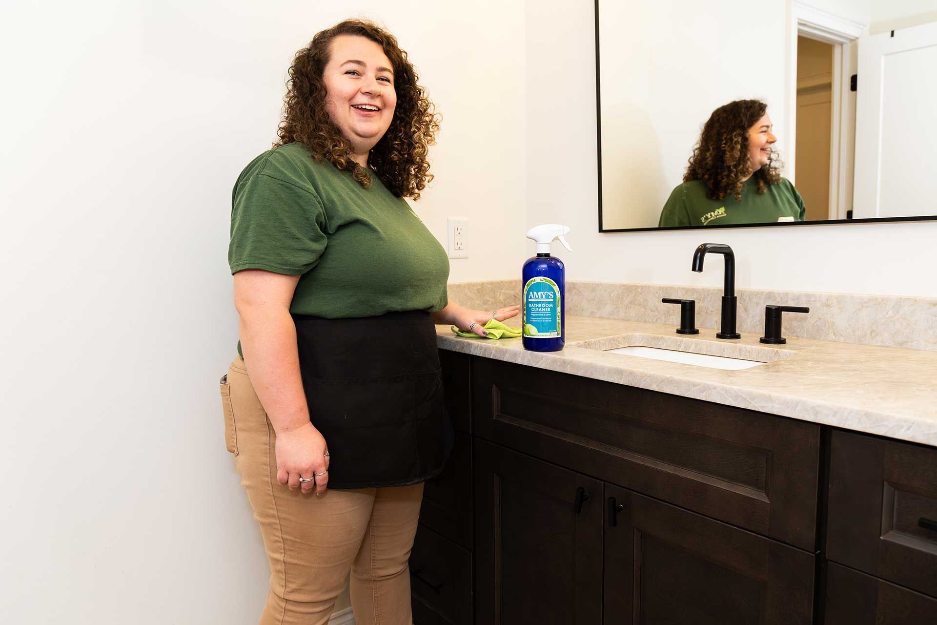 a woman in green shirt and apron smiles in a bathroom, holding a cleaning product