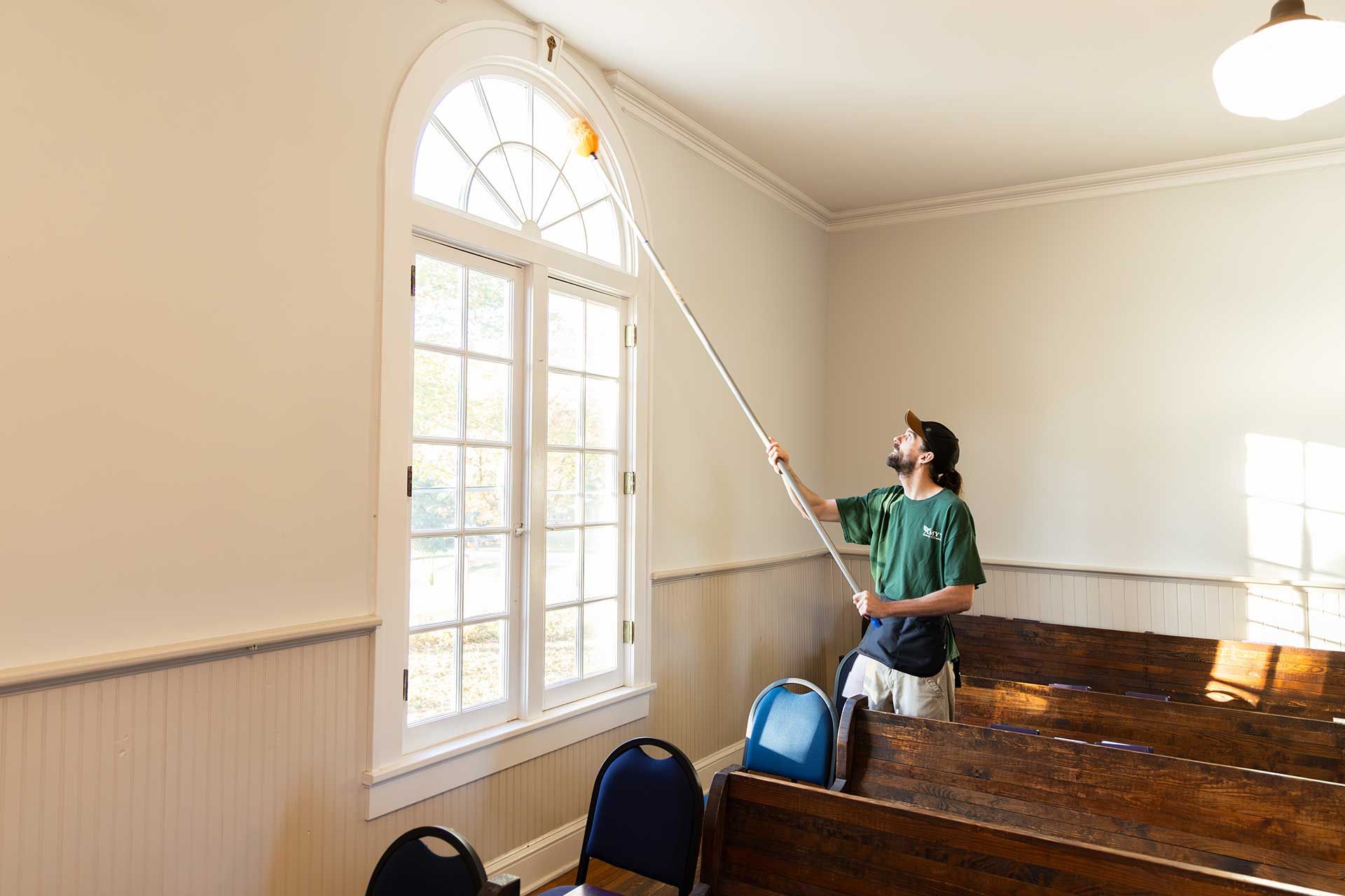 a person cleaning a high window with an extendable duster inside a room with a tall window