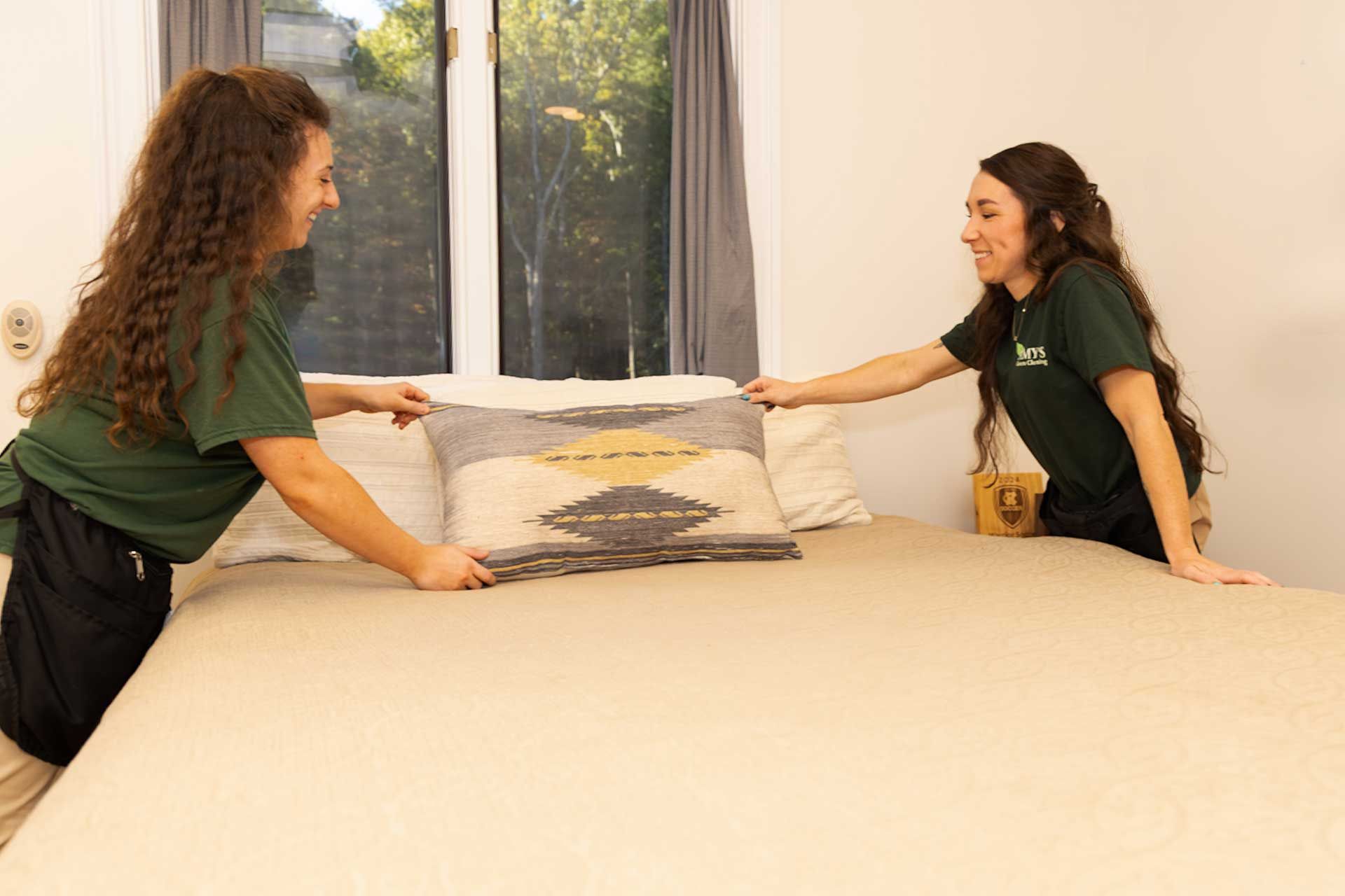 two people making a bed in a room with a window, smiling and holding a decorative pillow