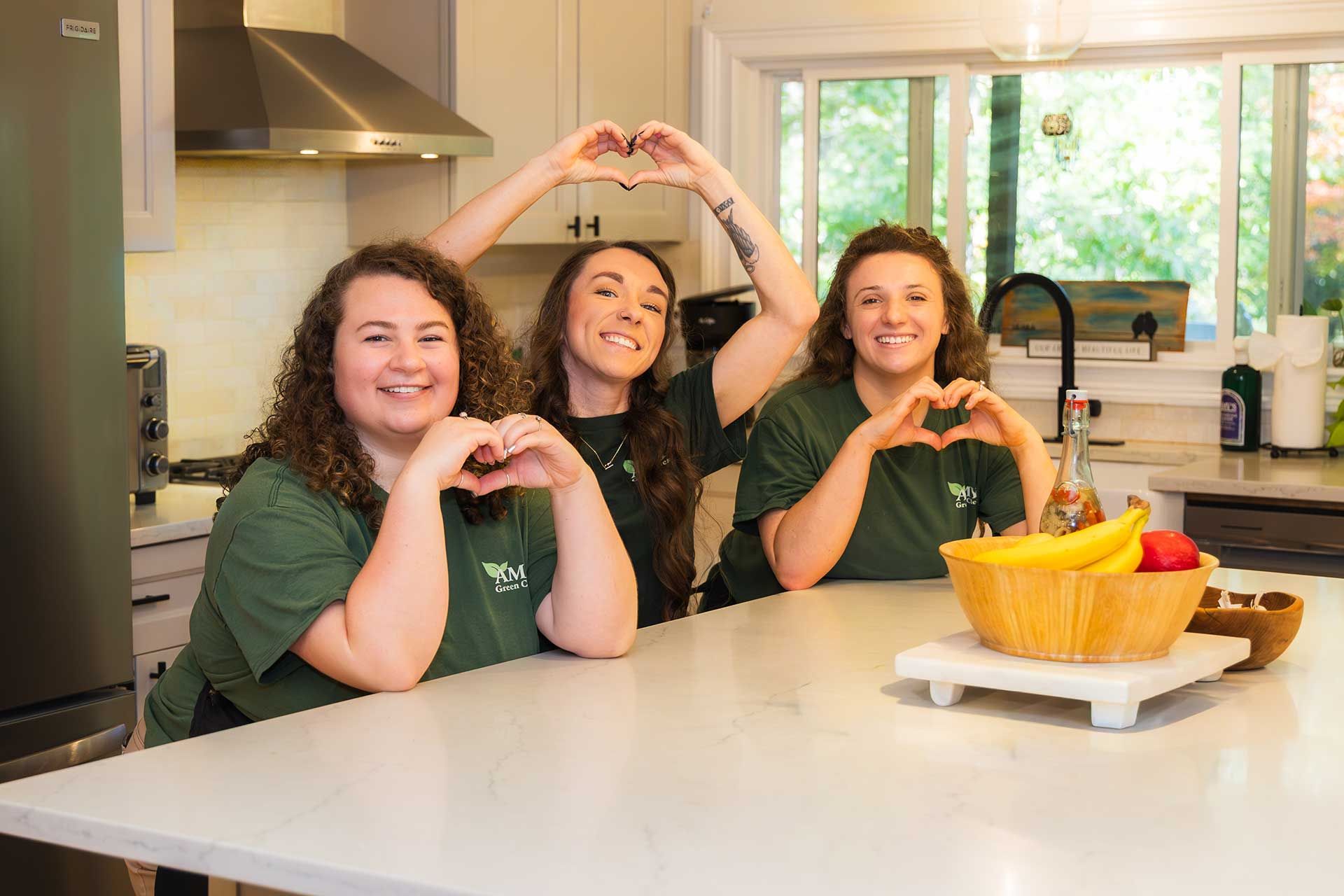 three people in green shirts make heart shapes with their hands in a kitchen