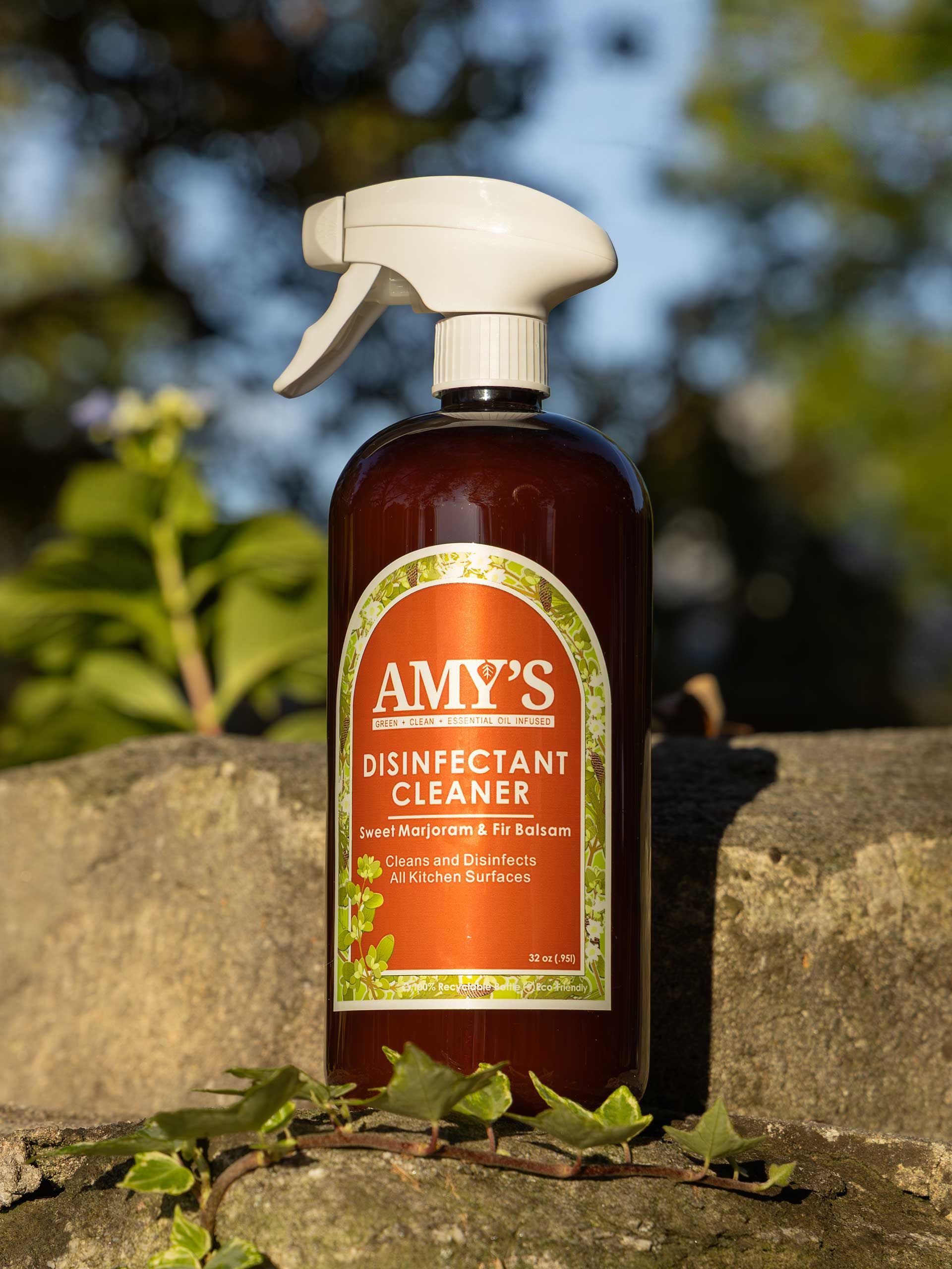 a bottle of Amy's Disinfectant Cleaner with white spray nozzle, on a stone surface with greenery in the background