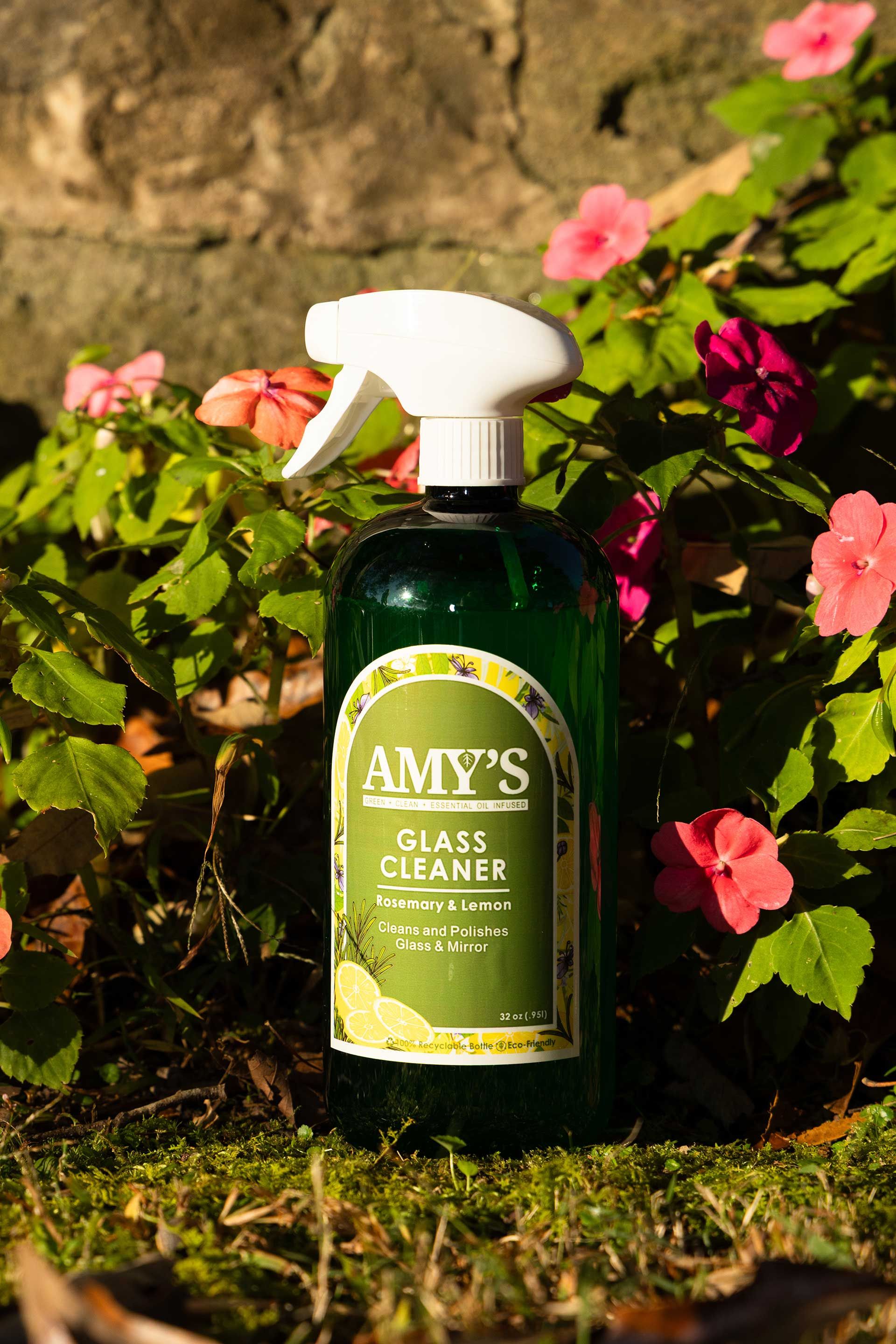 a bottle of Amy's glass cleaner with a white spray nozzle, set amidst green foliage and pink flowers