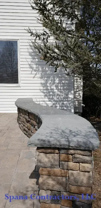 Stone bench with a concrete top next to a white house with a tree casting shadows.