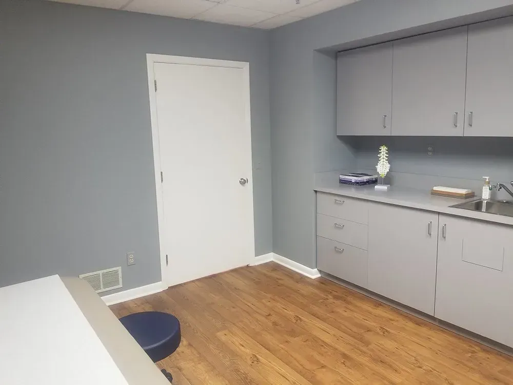 A medical exam room with gray cabinets, a sink, a door, and wood flooring.