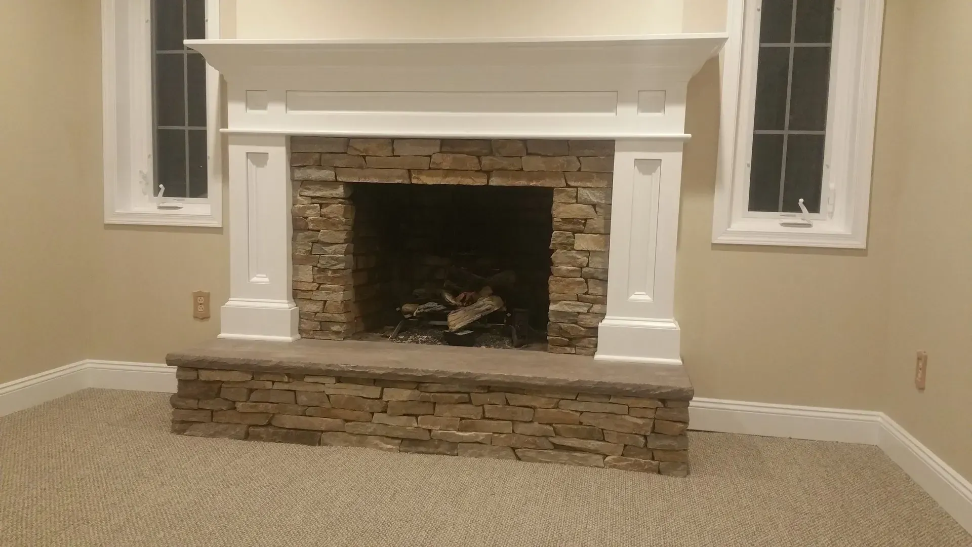 Fireplace with stone surround, white mantel, two windows on either side, beige walls, and carpeted floor.