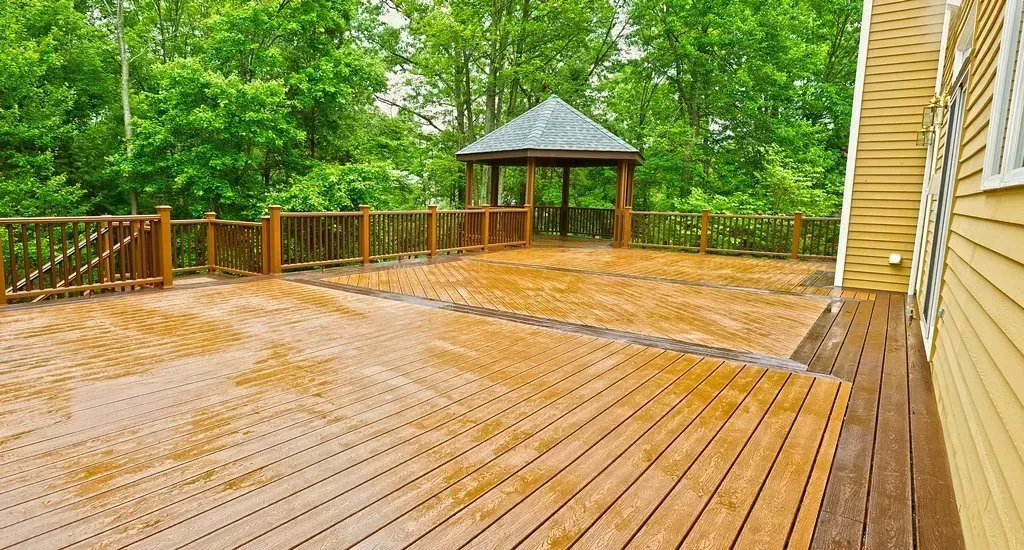 Wooden deck with a gazebo, surrounded by trees, and a yellow house siding.