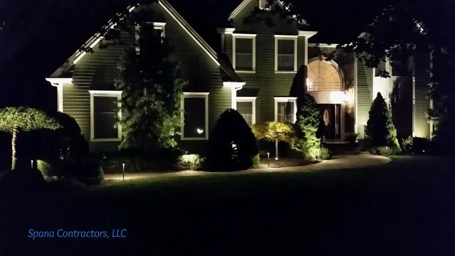 House at night with landscape lighting. Green exterior, white trim, and illuminated features.