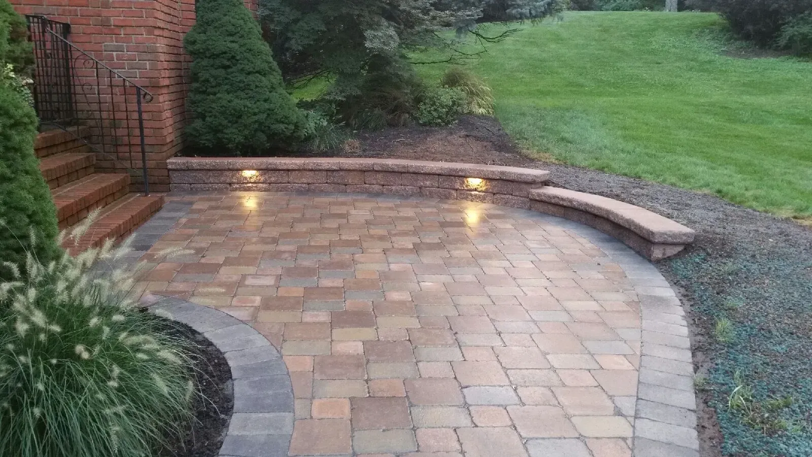 Brick patio with curved bench, lit by wall-mounted lights, next to a grassy area and brick house.