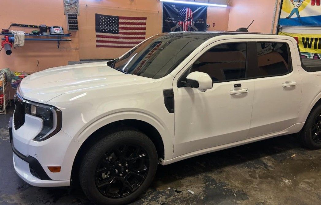 White Ford Maverick pickup truck with black accents parked inside a garage; American flag in the background.